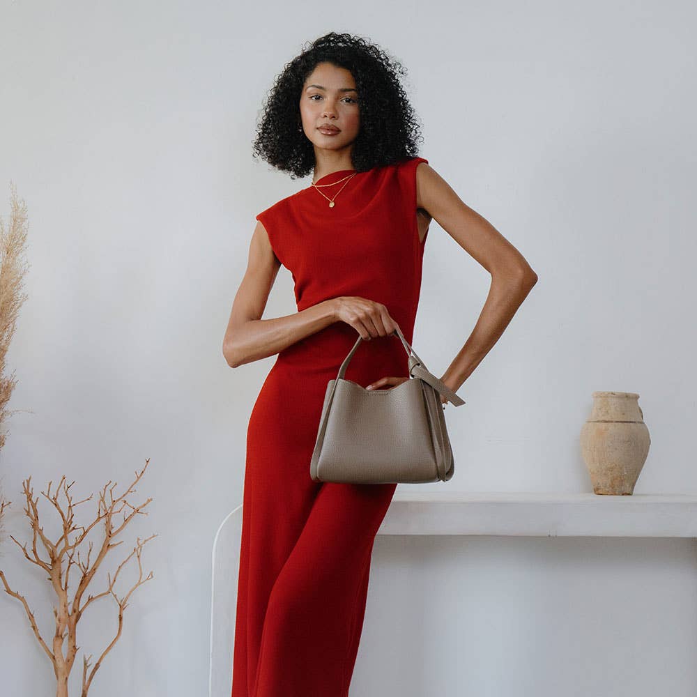 A woman with curly hair in a sleeveless red dress poses with the Felicity Red Recycled Vegan Top Handle Bag. She stands against a minimalist white background, near a dry branch in a vase and an earthenware pot on a shelf, looking at the camera with one hand on her hip.