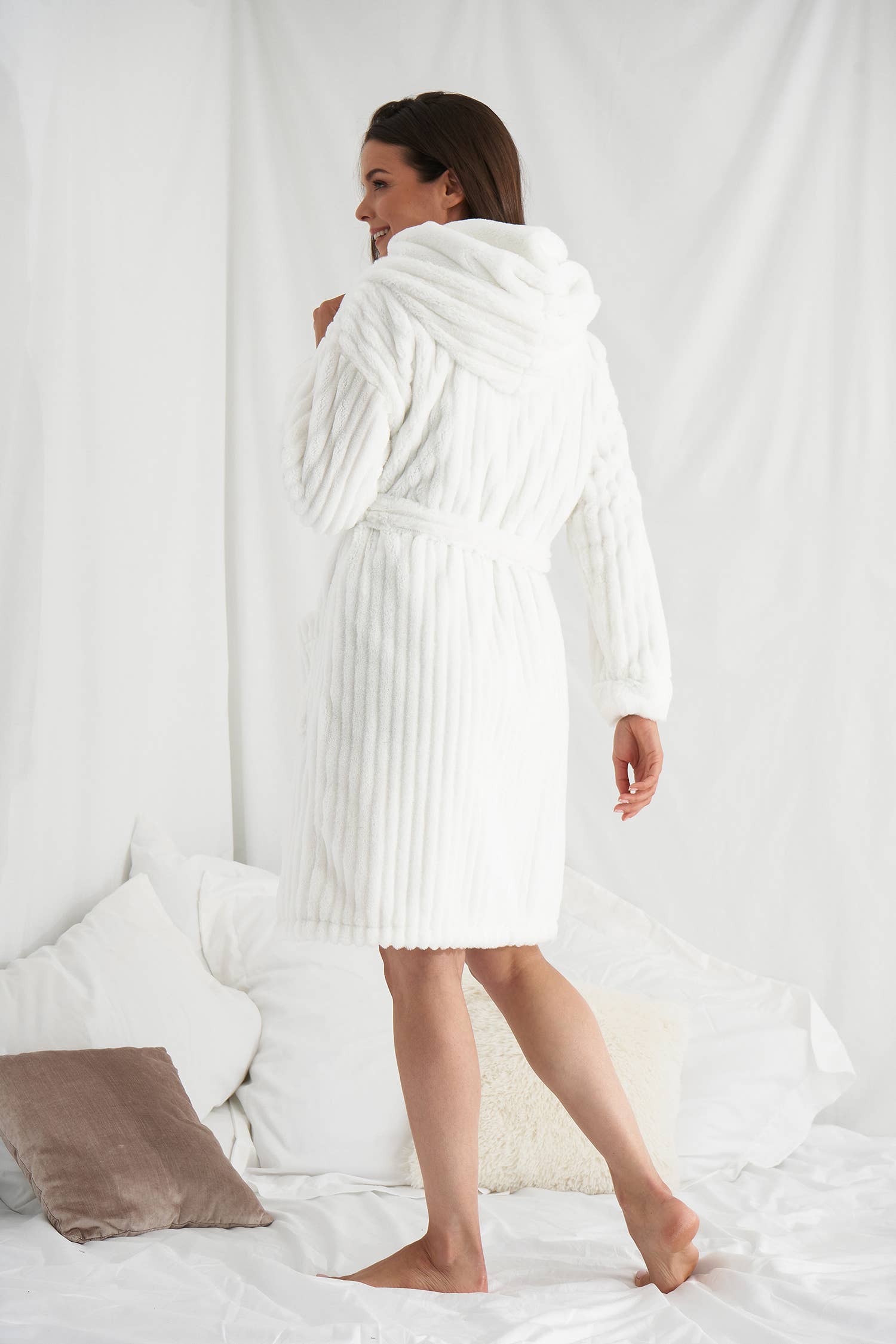Standing on a bed adorned with white sheets and pillows, a person with long hair is enveloped in the luxurious Cloud Robe, complete with a hood and belt. They smile while looking to the side, surrounded by the serene backdrop of white drapes.