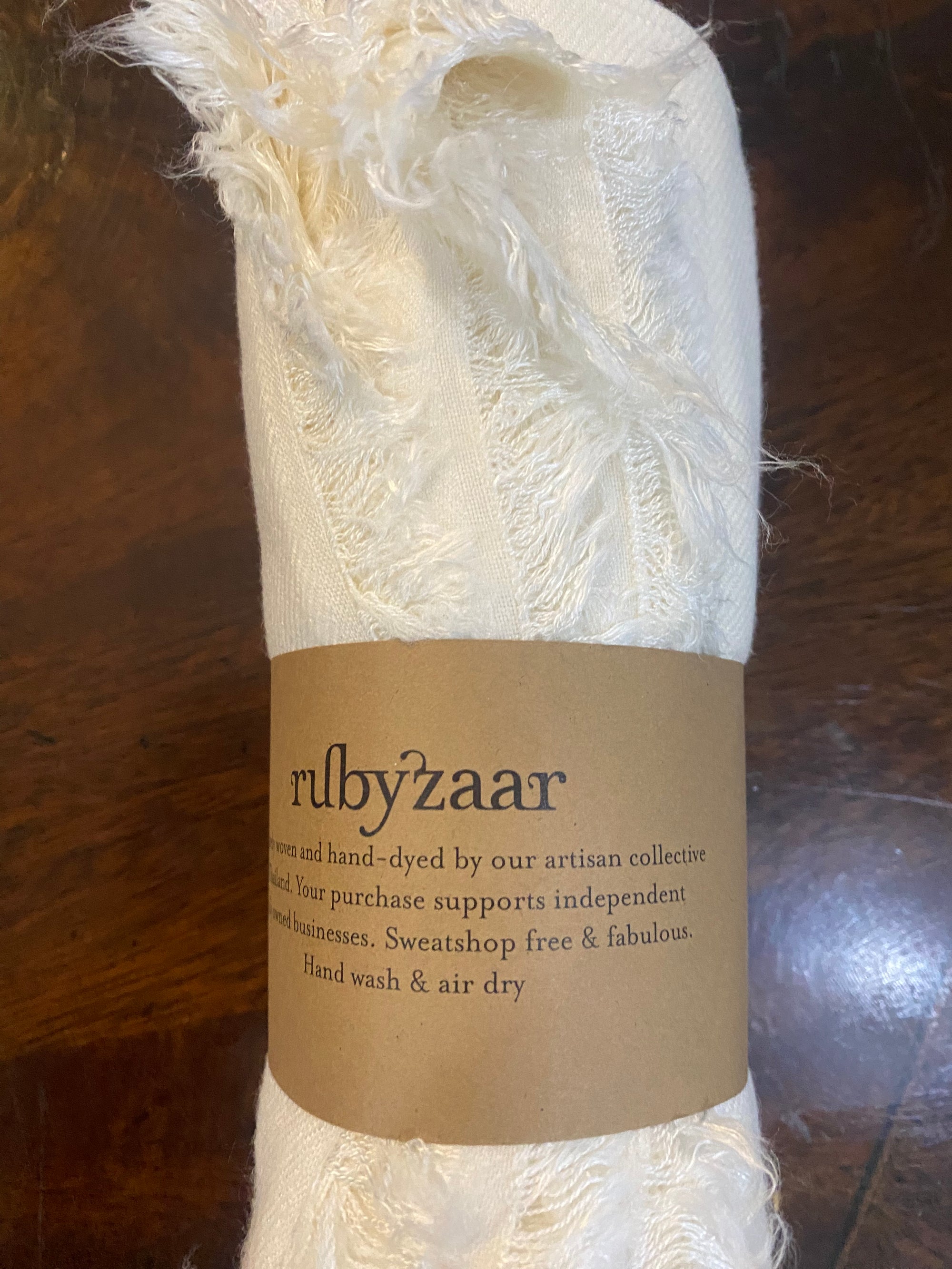 The Hima Large Scarf, Wrap, Shawl is a cream-colored, hand-woven accessory elegantly rolled with a brown paper label bearing the name "rubyzaar." Emphasizing its fair trade credentials, this product supports independent artisans. The scarf is hand-dyed for sustainable fashion and comes with recommendations for hand washing and air drying. Its fabric features charming frayed edges.