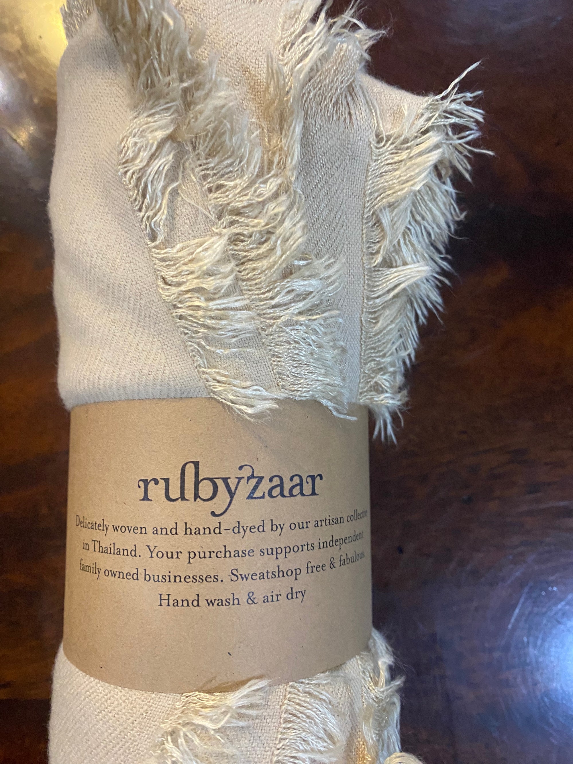 A close-up of a rolled, fringed Hima Large Scarf, Wrap, Shawl secured with a brown paper band labeled "rubyzaar," showcasing hand-woven pieces crafted in Thailand. This item embodies sustainable fashion and supports fair trade, benefiting independent family businesses. Instructions are to hand wash and air dry, set against a dark wooden surface.