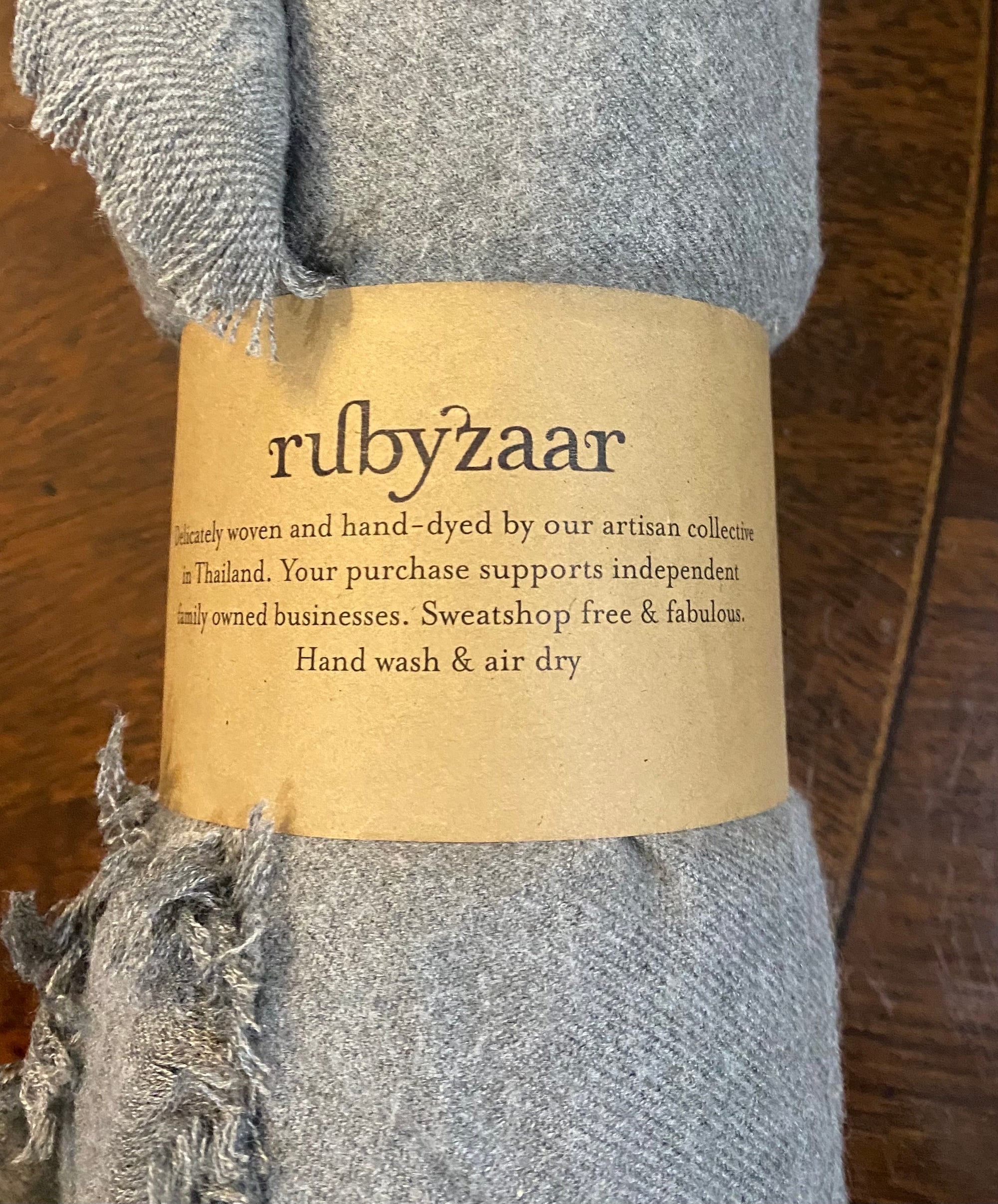 A roll of soft gray fabric with frayed edges, wrapped in a brown paper label that reads: "Hima Large Scarf, Wrap, Shawl by rubyzaar. Delicately woven and hand-dyed by our artisan collective in Thailand. Fair trade and sustainable fashion supporting family-owned businesses. Sweatshop free & fabulous. Hand wash & air dry.