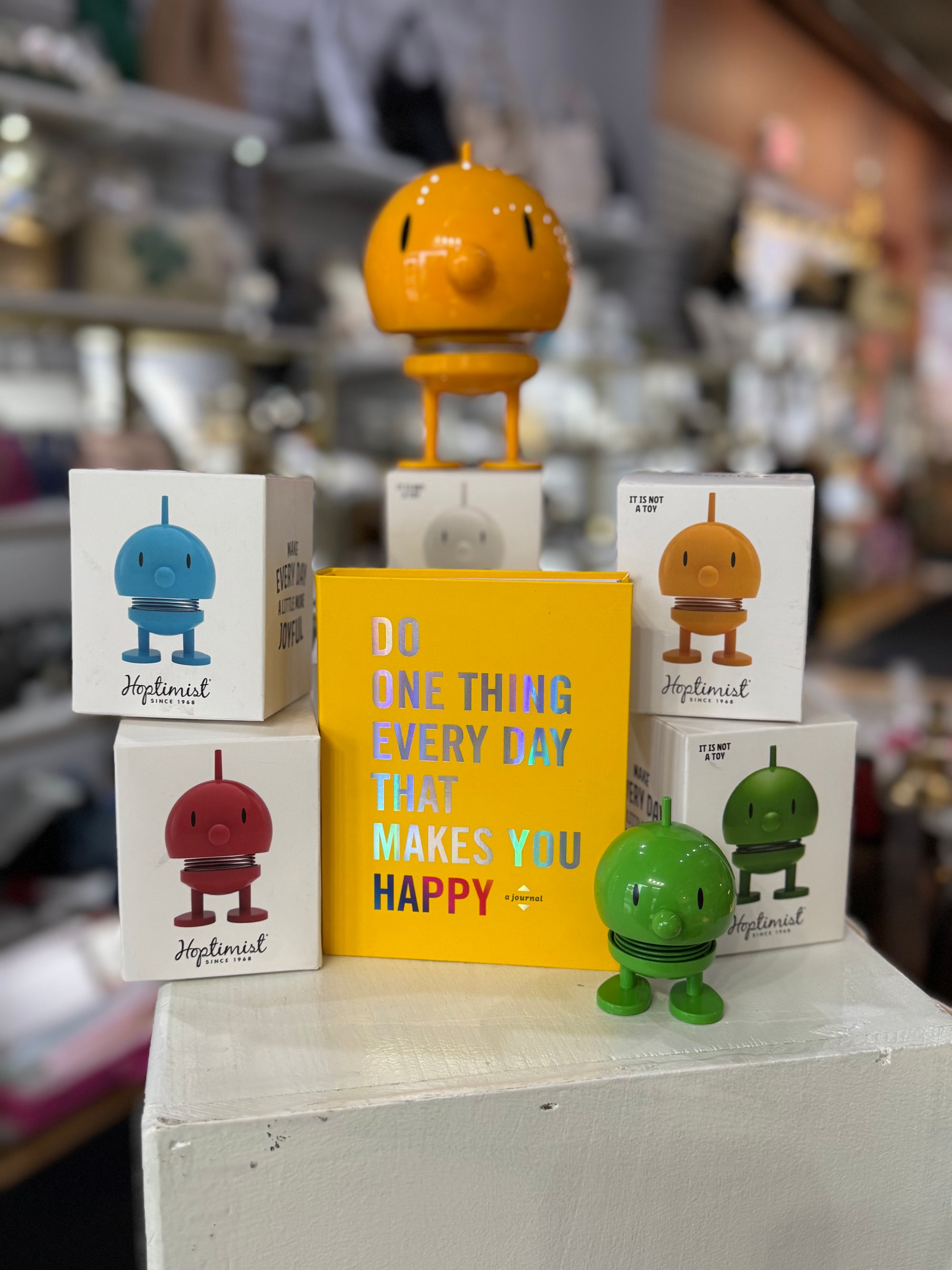 A display showcases "The Happiest Gift Set for Any Occasion" with Hoptimist figures in orange, blue, red, and green spreading joy. An orange figure on a book stack joins a yellow one promoting happiness. Shelves filled with items softly blur in the background.
