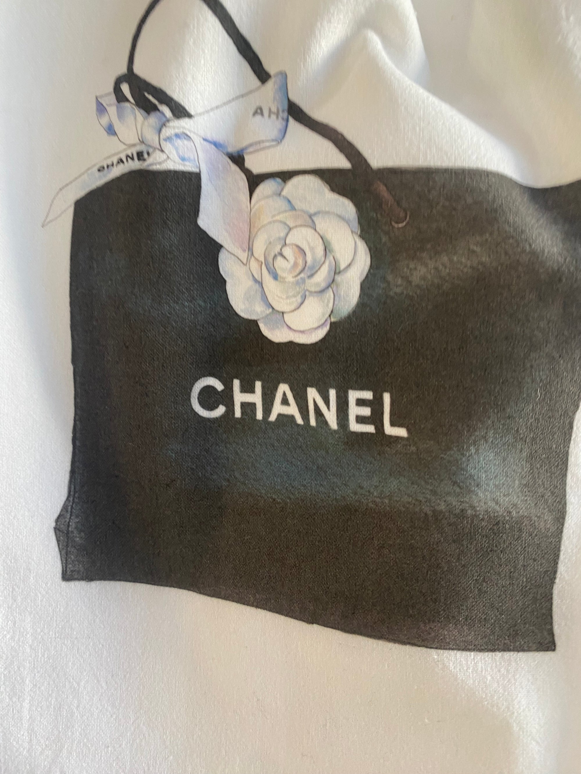 The fabric showcases a Chanel shopping bag in black, custom designed with white handles and a ribbon. It prominently features the iconic Chanel logo in white letters and is adorned with a handcrafted white camellia flower. The background is textured in light hues. This design is found on the Tea Towels - Chanel product.
