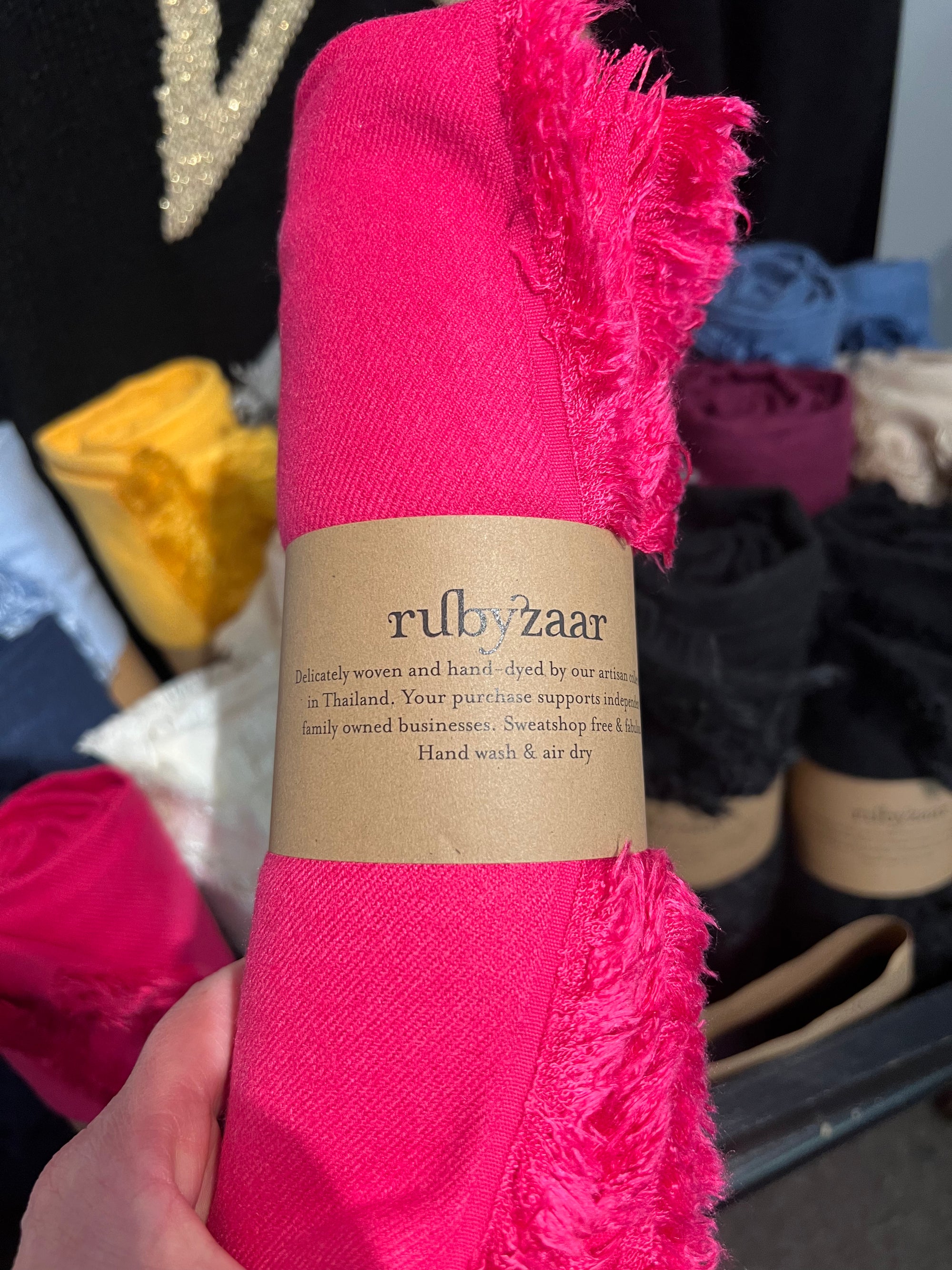 A hand presents a vivid pink woven Hima Large Scarf, Wrap, Shawl with frayed edges. It is adorned with a brown label that displays "rubyzaar" and emphasizes its fair trade origins. The label also notes the hand-dyed nature and Thai craftsmanship of the product. In the background, more folded fabrics, including other handcrafted scarves, are visible in a bin.
