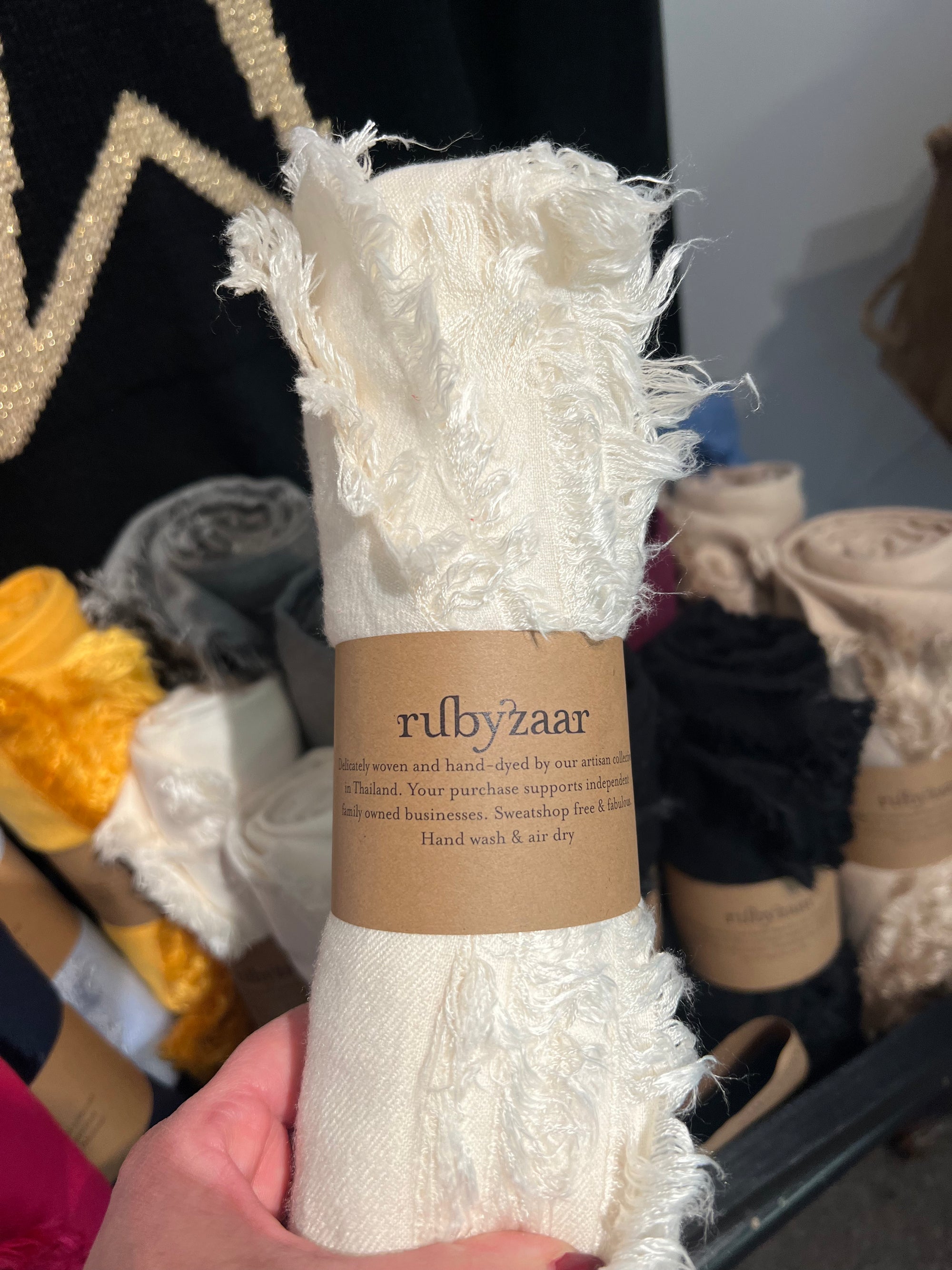 Close-up of a hand holding the Hima Large Scarf, Wrap, Shawl, a cream-colored textile with frayed edges. The label reads "rubyzaar" and indicates it is handmade and dyed by artisans in Thailand. This fair trade piece exemplifies sustainable fashion, with surrounding rolls in various colors blurred in the background.