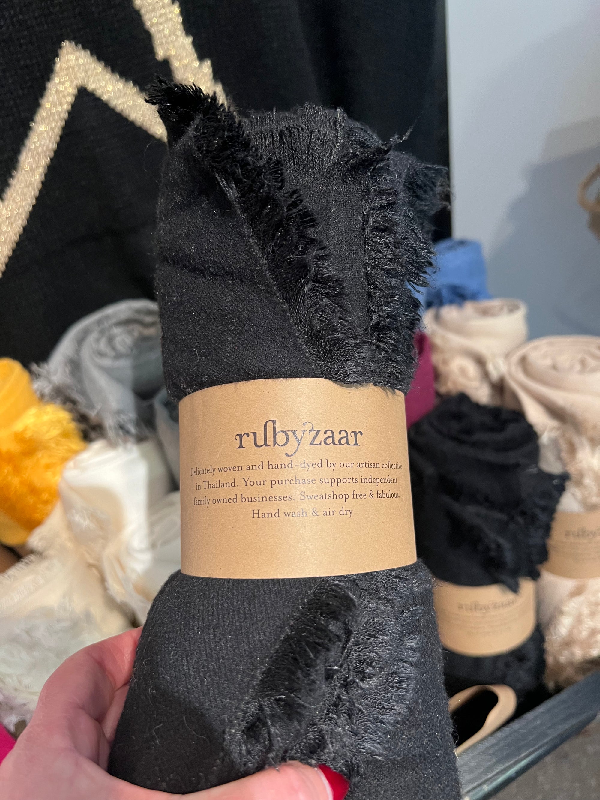 A close-up captures a hand holding a roll of black fabric with frayed edges, symbolizing sustainable fashion. The fabric is part of the "Hima Large Scarf, Wrap, Shawl" collection by rubyzaar, suggesting its fair trade origins. Behind it, various rolls of colored fabrics are visible in shades of white, cream, yellow, and blue.