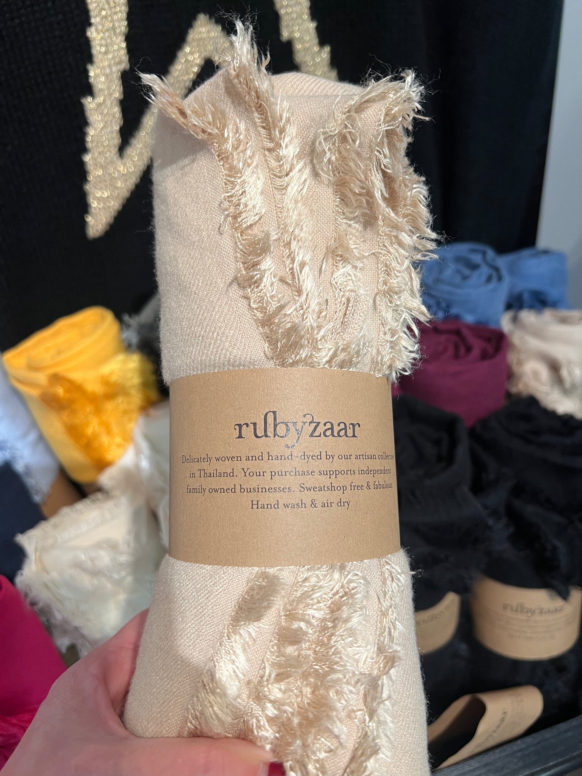A hand holds a rolled-up, fringed Hima Large Scarf, Wrap, Shawl with a "rubyzzaar" label. Behind it are more scarves in colors such as yellow, pink, and black. The label highlights that these fair trade pieces are handmade and naturally dyed by artisans in Thailand and suggests hand washing and air drying for best care.