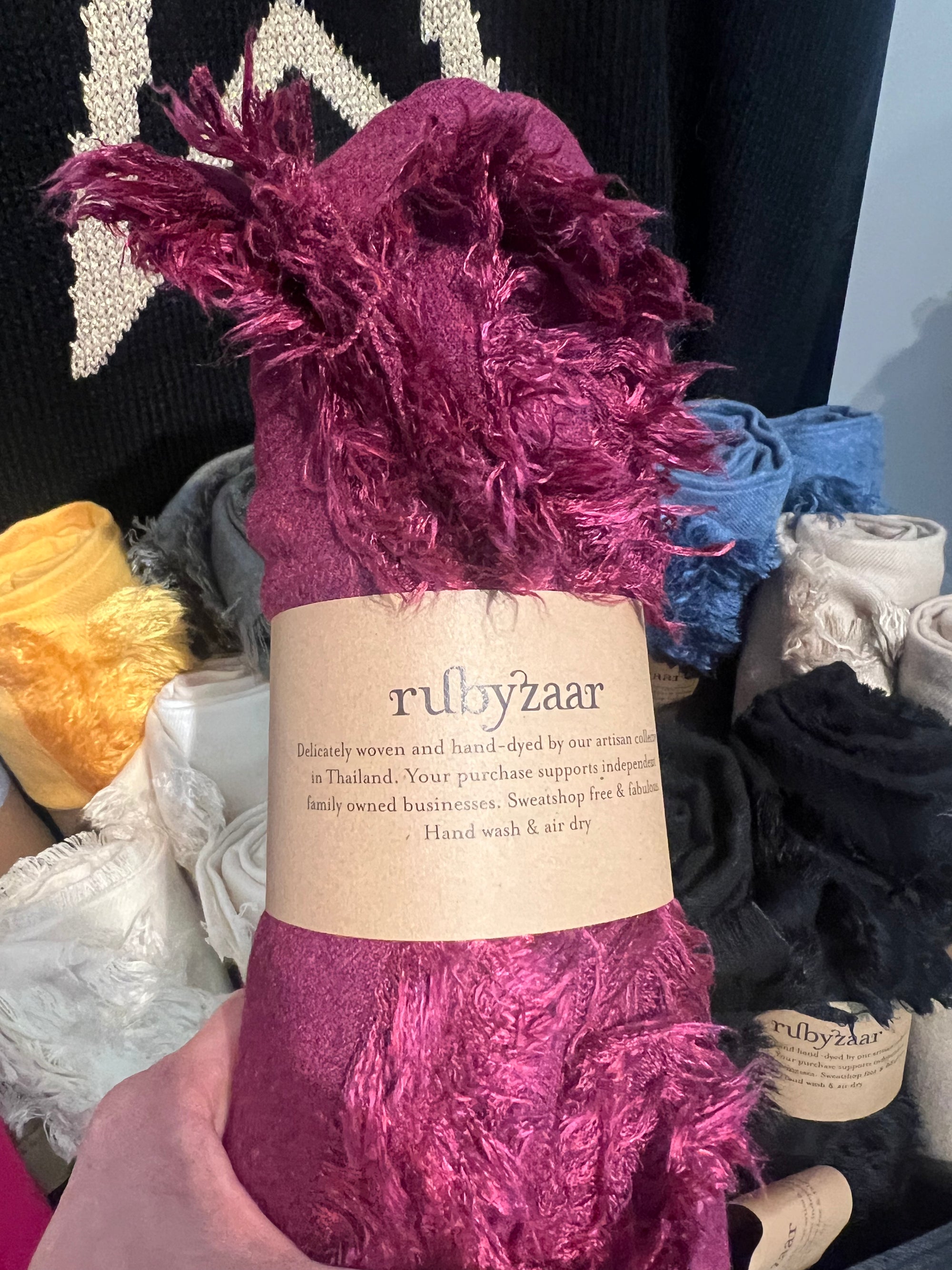 A close-up shot features a hand holding a deep pink, fringed Hima Large Scarf, Wrap, Shawl with a "rubyzaar" label. The label highlights the scarf as delicately woven and hand-dyed in Thailand, advocating for fair trade and sustainable fashion. Surrounding the centerpiece are rolls of similar hand-woven scarves available in various colors like yellow, white, and blue.