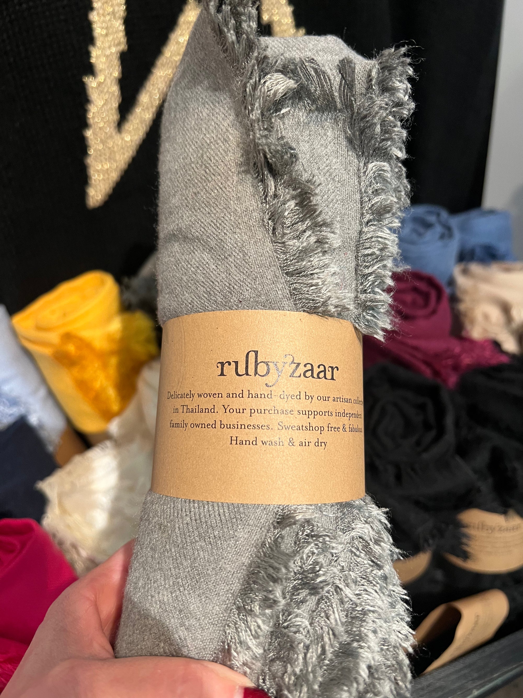A hand displays a fringed, rolled-up gray Hima Large Scarf with a brown "rubyzaar" label. The vibrant backdrop features an array of colorful textiles in yellow, blue, and red. Emphasizing the handmade allure of these fair trade, hand-woven scarves, every purchase supports indigenous businesses in Thailand through sustainable fashion.