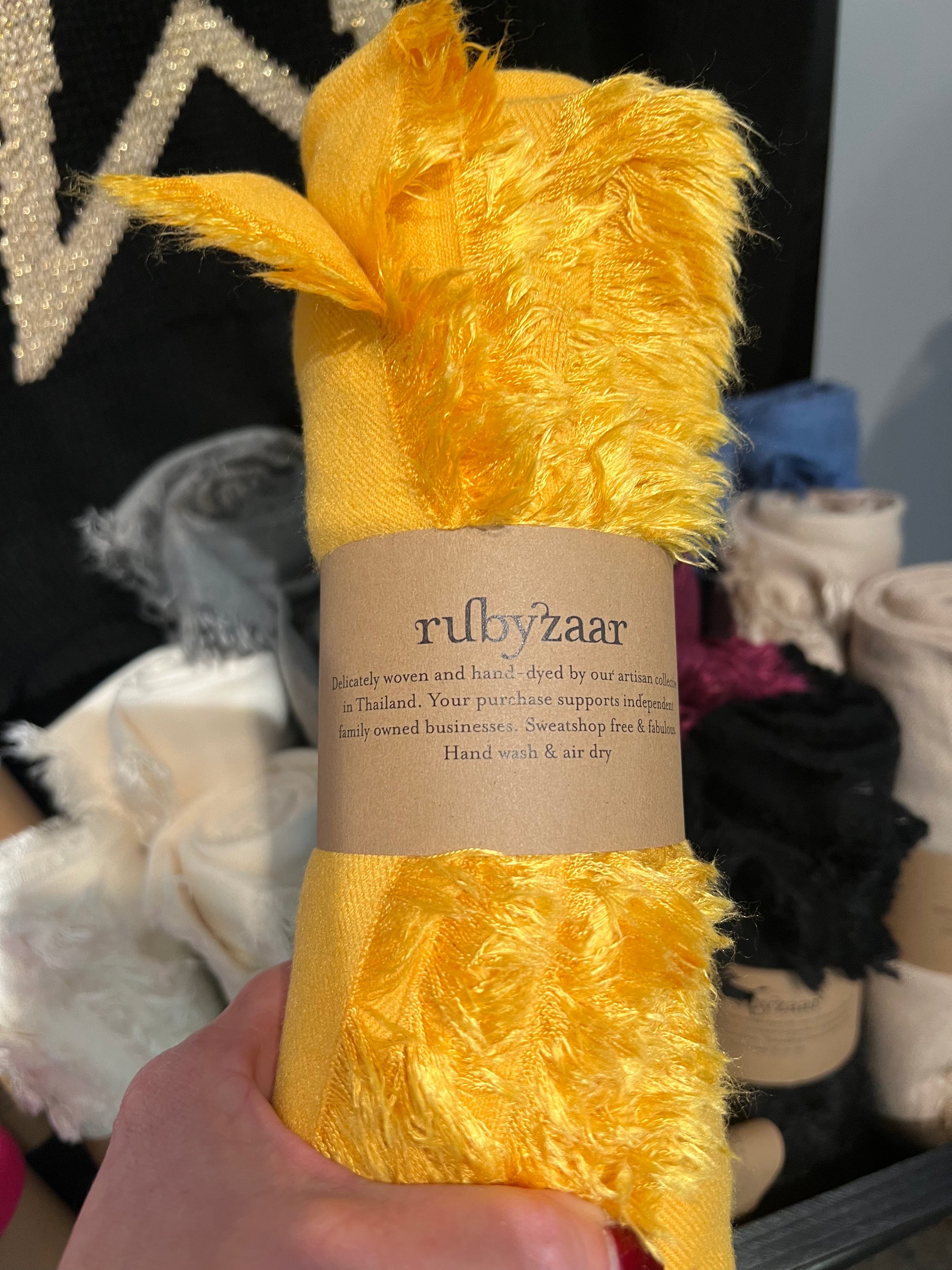 A hand holds a Hima Large Scarf, Wrap, Shawl, in bright yellow with a brown paper label that reads "rubyzaar." This soft, fuzzy textile is part of our sustainable fashion collection. Blurred in the background are multiple rolls of fair trade fabrics in an array of colors.