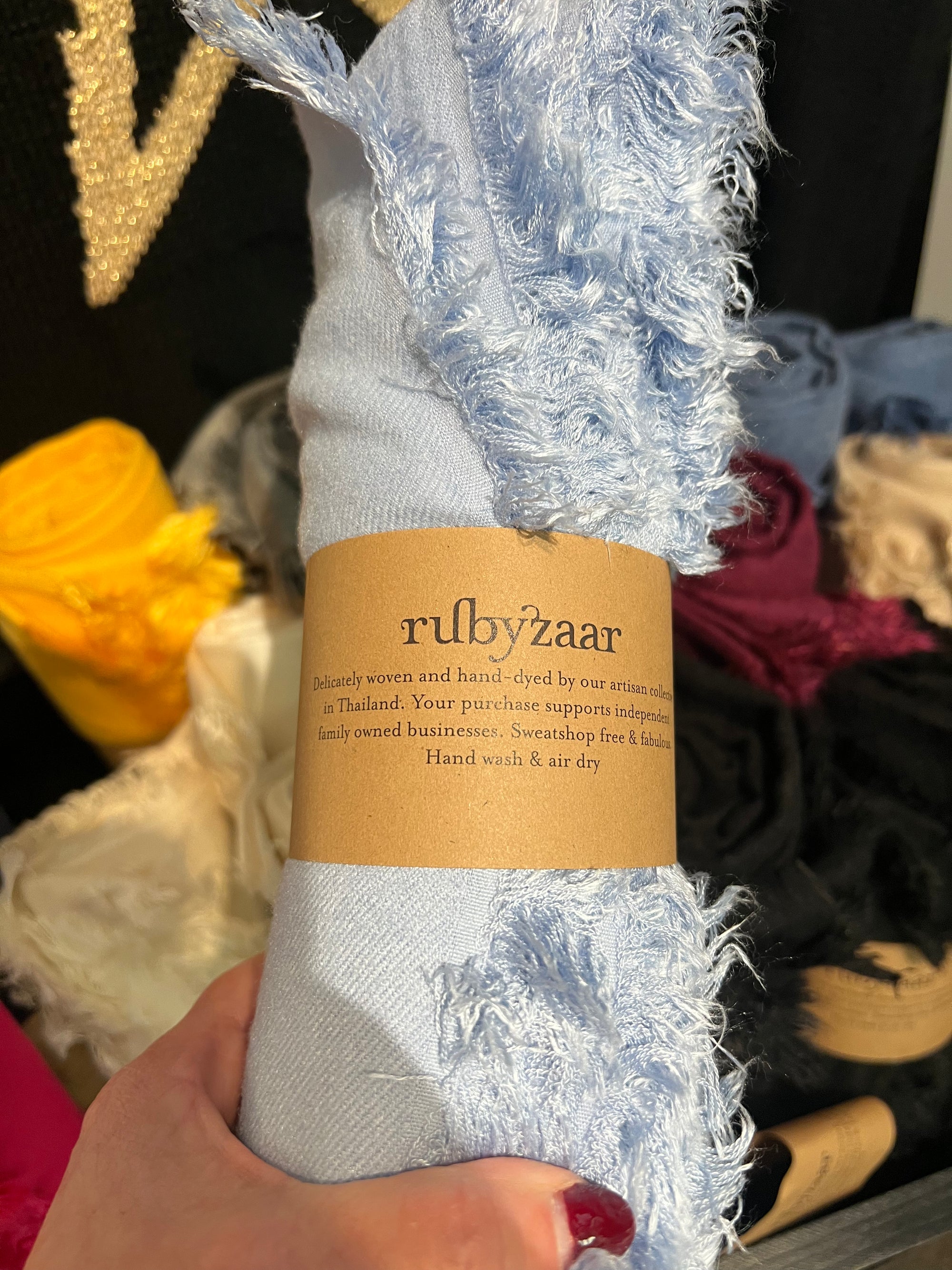 A hand holds a soft blue fringed fabric roll with a brown label that reads "Hima Large Scarf, Wrap, Shawl," highlighting its hand-woven, fair trade craftsmanship from Thailand. In the background, more vibrant rolls promise a colorful touch to sustainable fashion. The setting is an artisanal display area.