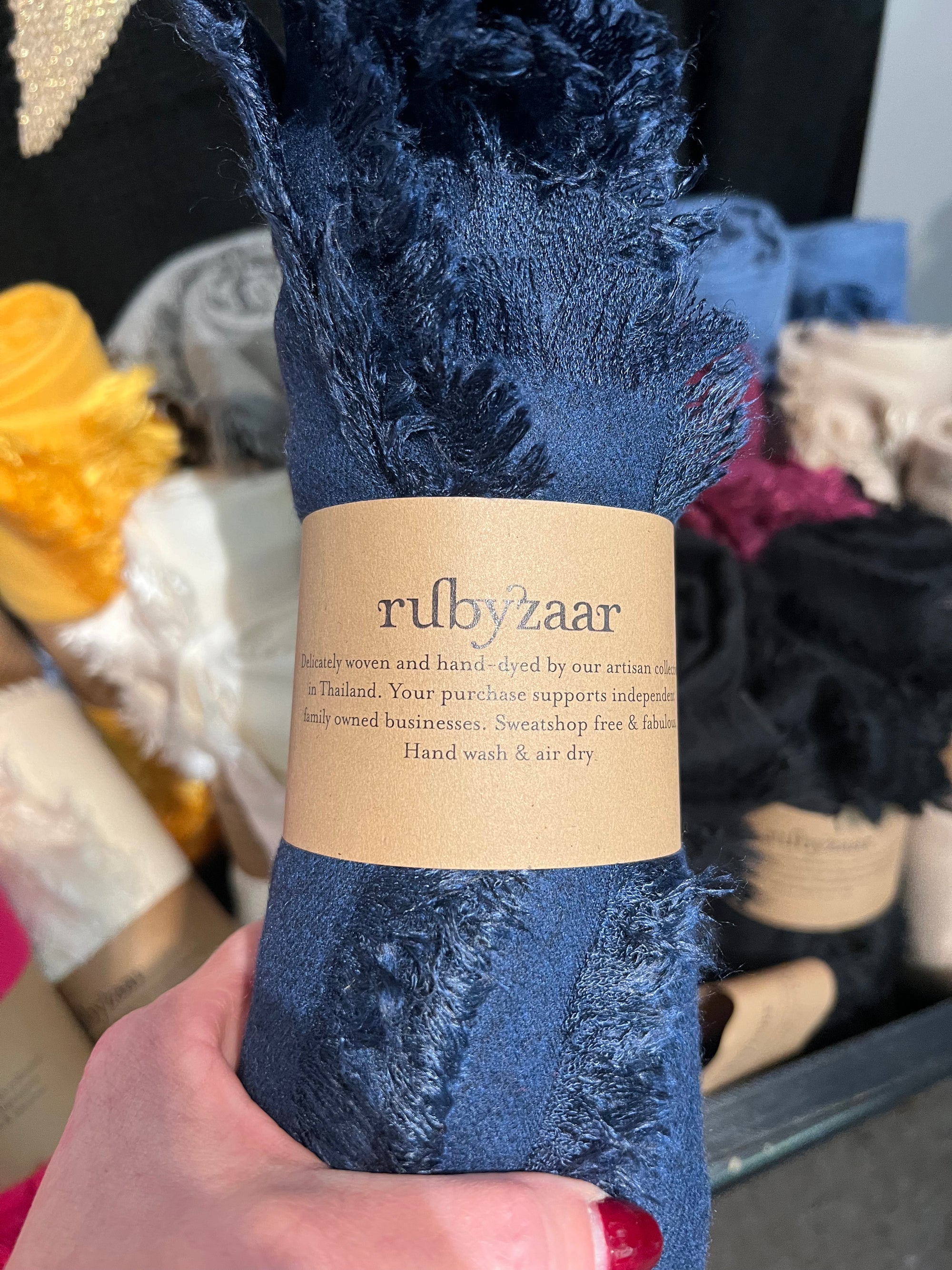 Close-up of a hand holding a Hima Large Scarf, Wrap, Shawl in deep blue with frayed edges. The label reads "rubyzaar," showcasing it as handmade and dyed in Thailand, supporting independent businesses through sustainable fashion. In the background, other fabric rolls in colors such as yellow, white, and black are visible.