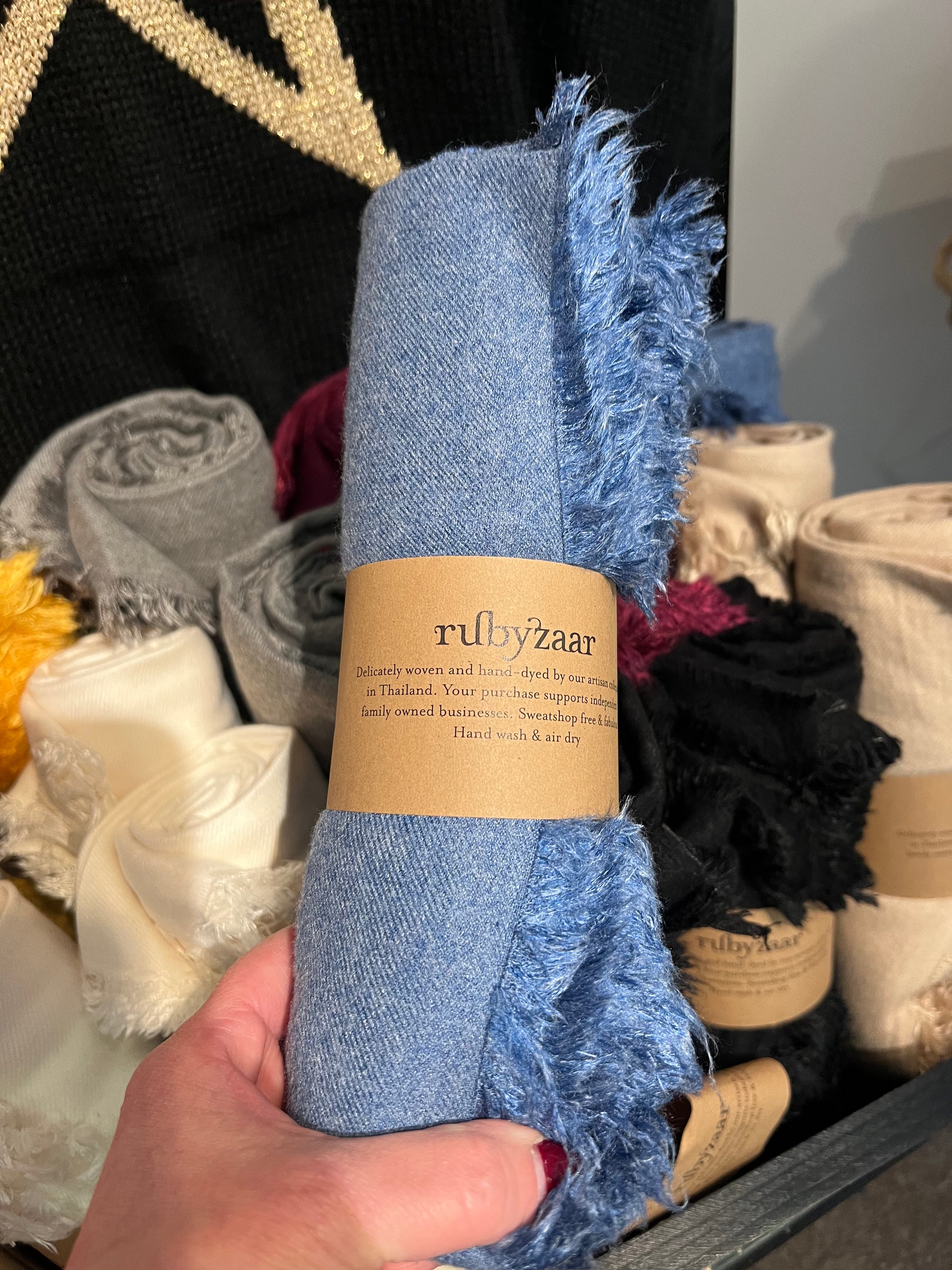 A hand holds a rolled-up Hima Large Scarf, Wrap, Shawl with fringed edges, wrapped in a brown paper label that reads "rubyzaar," epitomizing sustainable fashion. Behind it are fair trade, hand-woven scarves in various colors, neatly displayed in a box. The setting appears to be a retail environment.