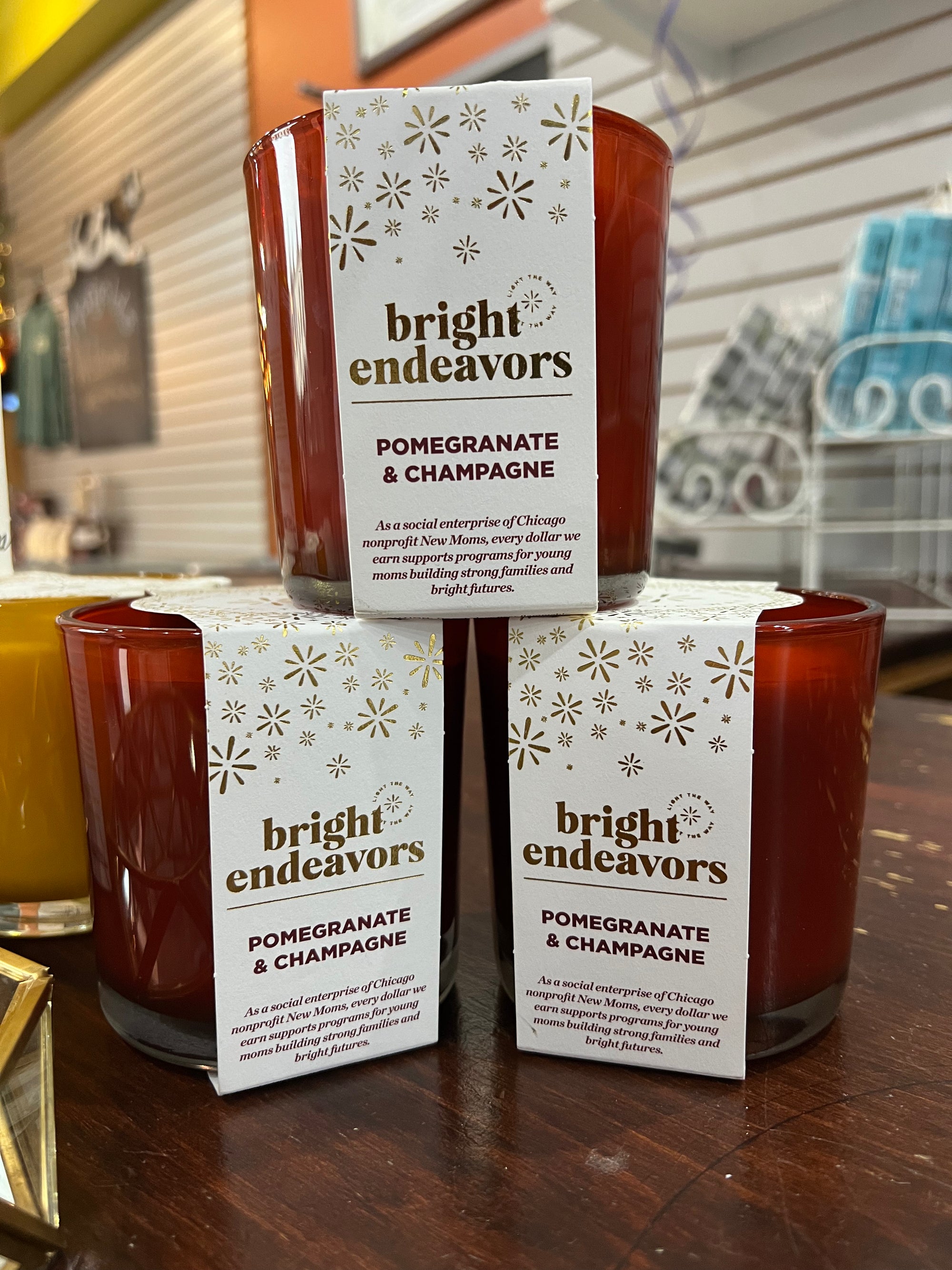Three "Pomegranate & Champagne Soy Candles," made from soy wax and boasting a phthalate-free fragrance, fill the cozy store with their red glow. Two candles sit at the bottom and one on top, all resting on a wooden surface against shelves filled with various items.