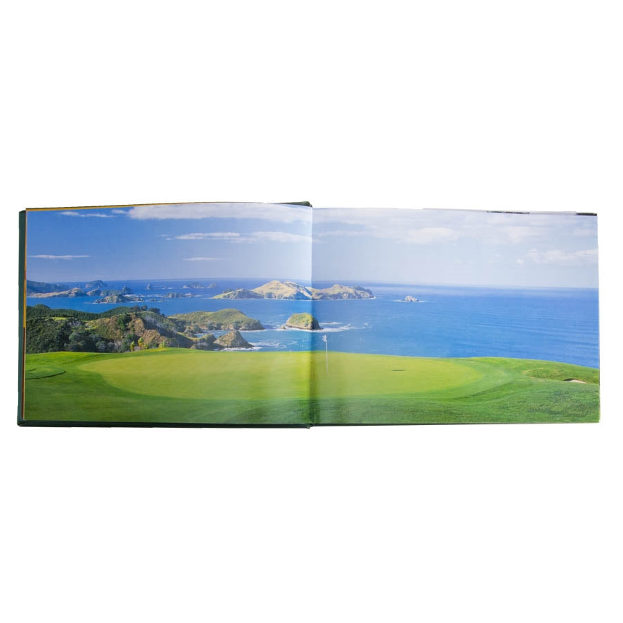 In the open pages of "Golf Courses - Fairways of the World - Leather Bound Edition," a stunning panoramic photograph by David Cannon is on display. The image captures a vibrant green field reminiscent of renowned golf courses, featuring dramatic cliffs that descend to a serene blue ocean under a clear sky. Small islands and rolling hills create a captivating panorama along the distant horizon.