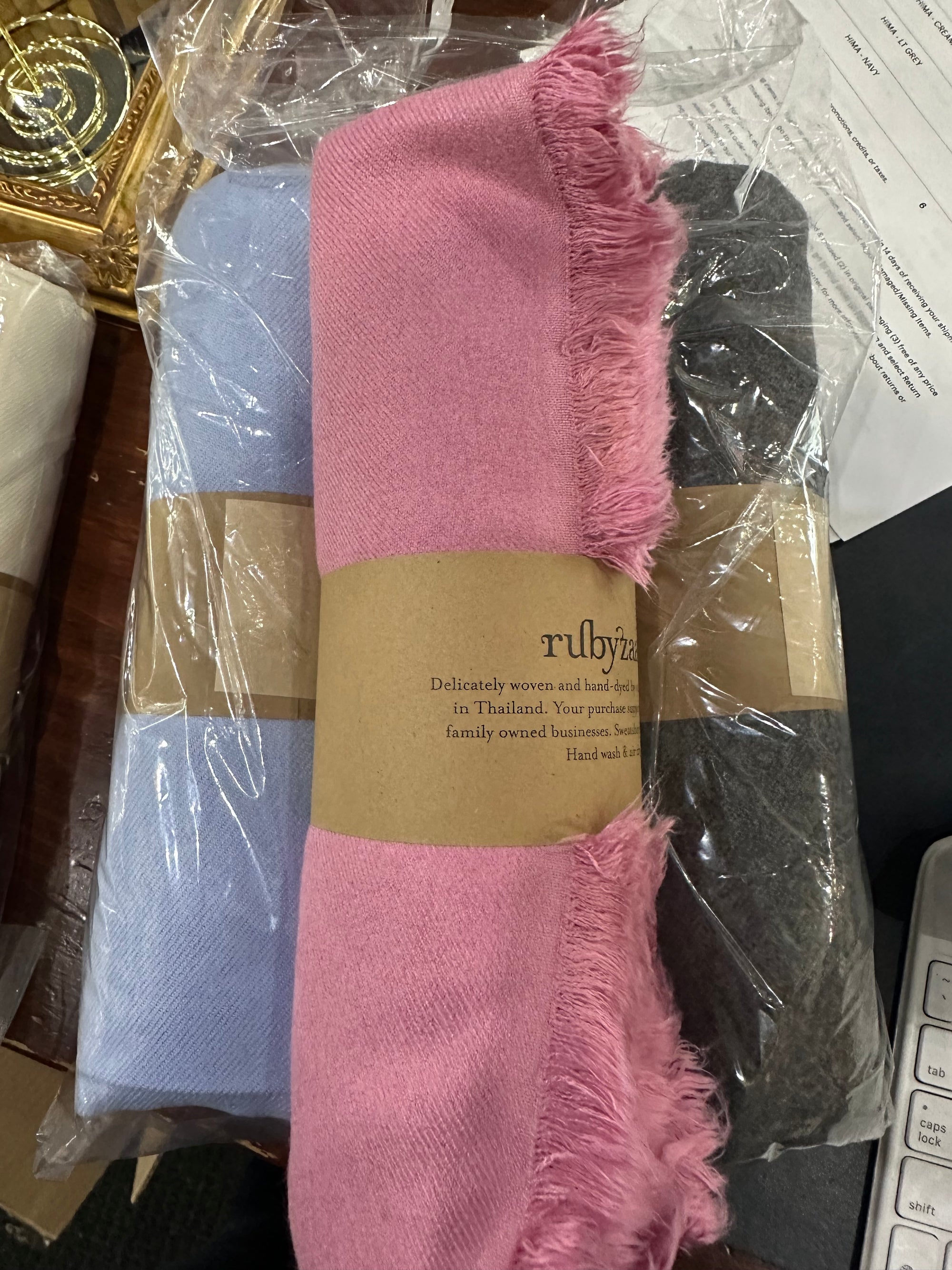 Three Hima Large Scarves in pink, blue, and gray are packaged in clear plastic. Each scarf features a brown paper band with text. These hand-woven scarves are displayed on a table next to a piece of paper and a small section of a keyboard.