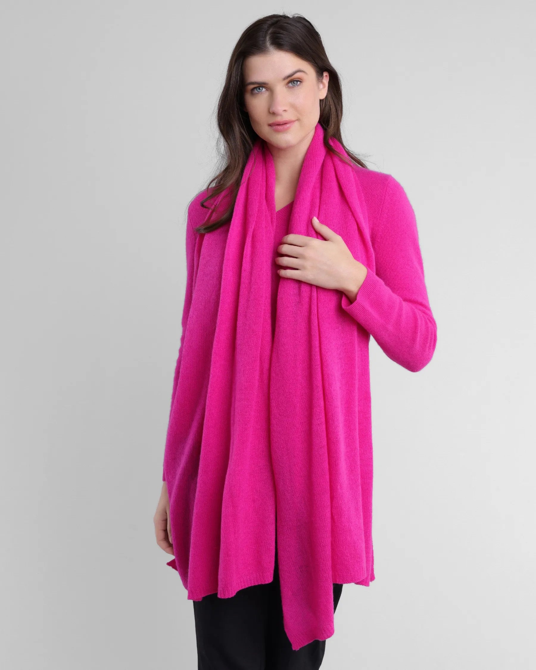 A woman with long dark hair wears the Cashmere Poncho in bright pink, paired seamlessly with a matching scarf. Her right hand delicately holds the scarf as she smiles softly, dressed in black pants against a light gray background.