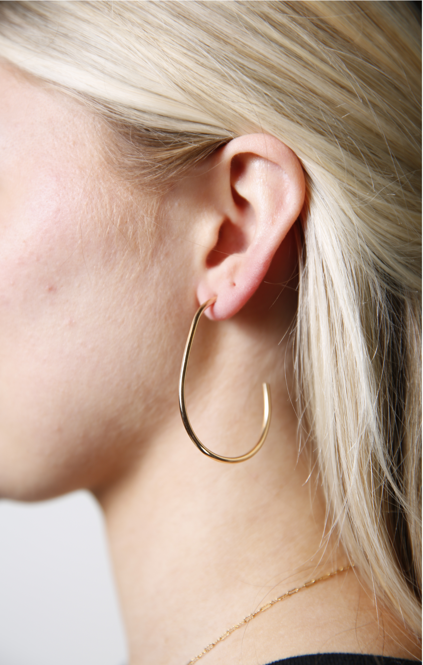 A close-up shows a person with light blonde hair wearing a large Pear Hoop earring made of 14k gold-filled material, elegantly adorning their left ear and complementing their light skin tone. A delicate gold necklace is partially visible against a black top, set against a soft, neutral background.