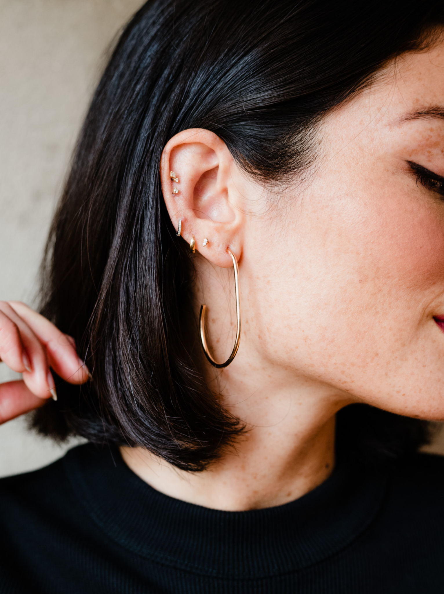 A woman with short black hair showcases handcrafted jewelry, wearing a pair of large 14k gold-filled Pear Hoops on her left ear along with multiple small studs. Dressed in a black top, she gently touches her hair with her right hand against a softly blurred background.