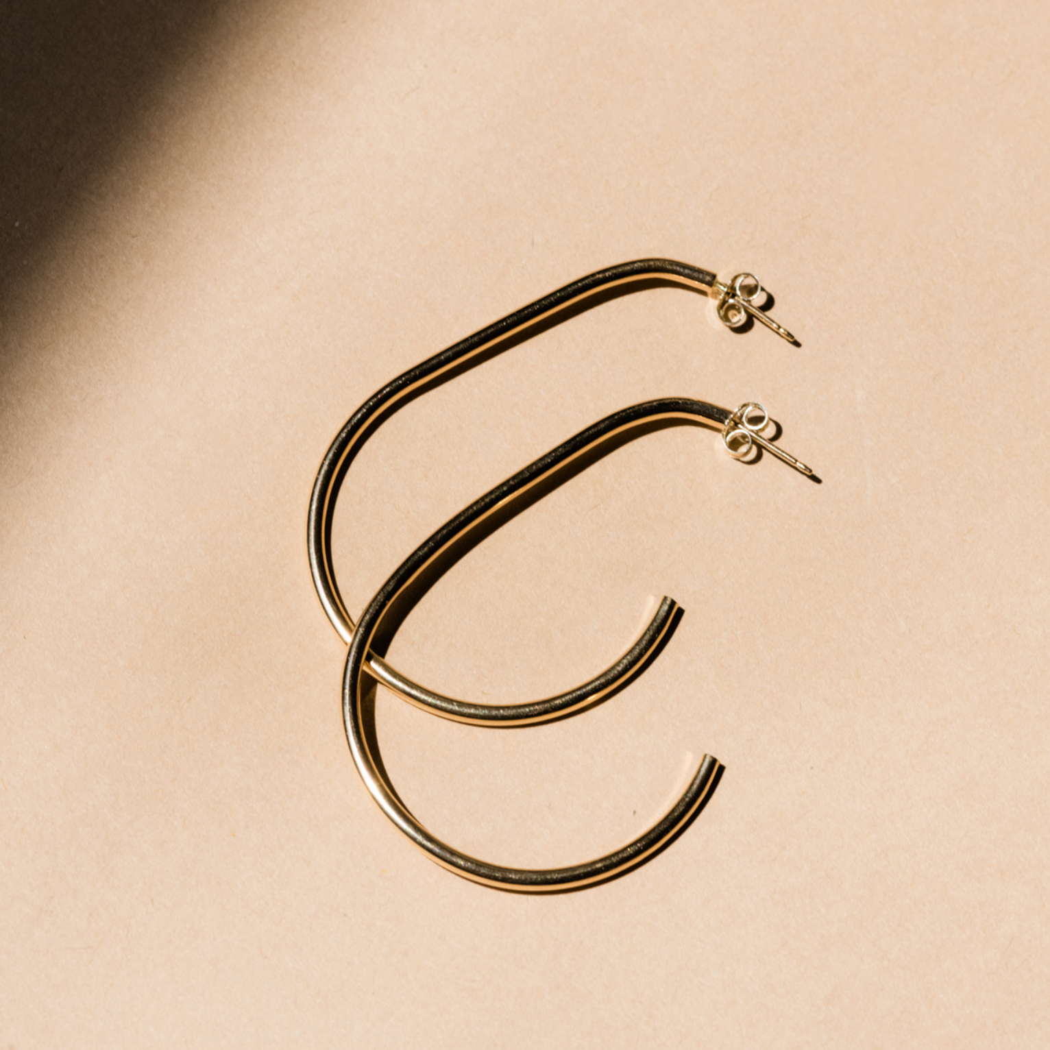 The Pear Hoops, a pair of large, curved hoop earrings crafted from 14k gold-filled material with post and butterfly backings, lie gracefully on a smooth beige surface. They cast soft shadows while a diagonal beam of light accentuates their shiny metallic texture.