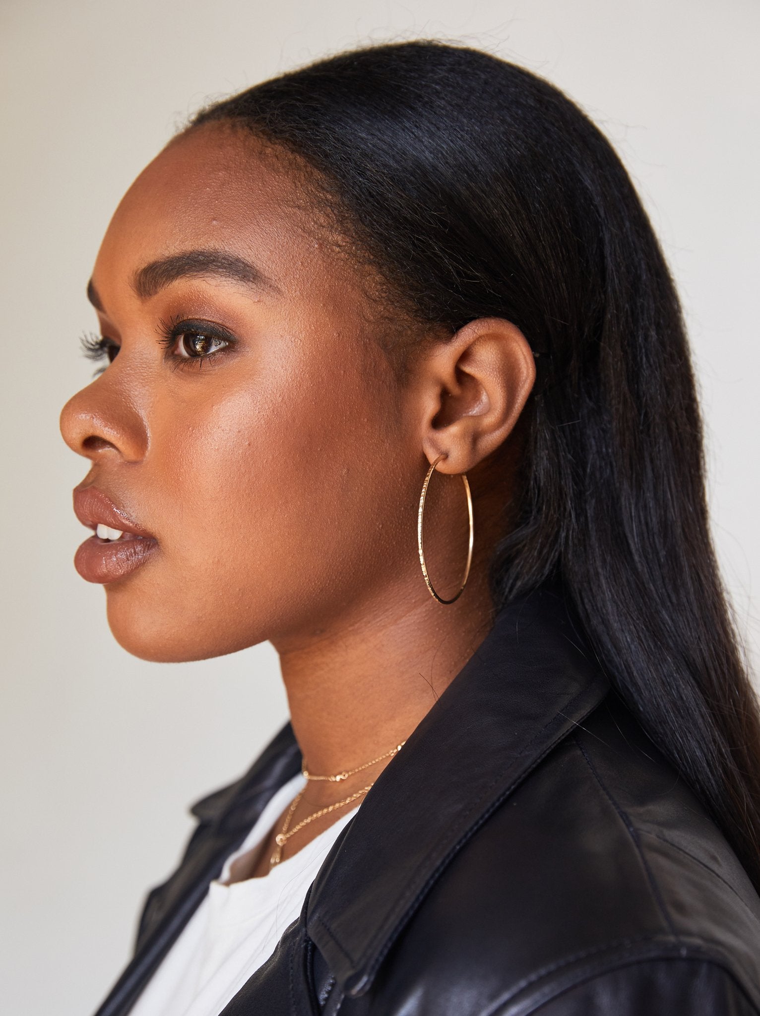 Profile view of a person with long black hair, wearing Luxe Hoops made of 14k gold fill and layered necklaces. They are dressed in a black leather jacket over a white top. The background is plain and light-colored.