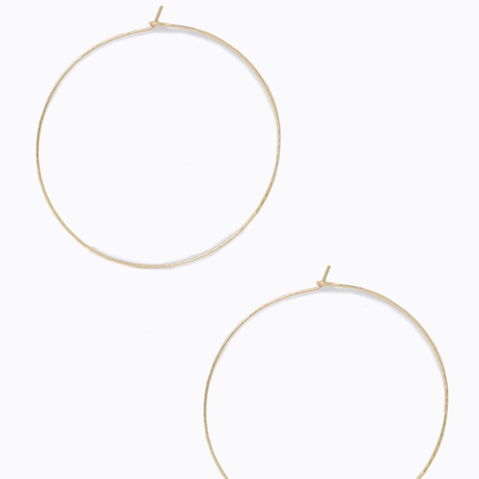 A pair of Luxe Hoops, crafted from 14k gold-fill, rests elegantly on a plain white background. These large hoop earrings boast a classic and minimalist design with their simple and unadorned appearance. Each thin hoop is equipped with a discreet clasp at the top for fastening, while their lustrous gold surface gently reflects light.