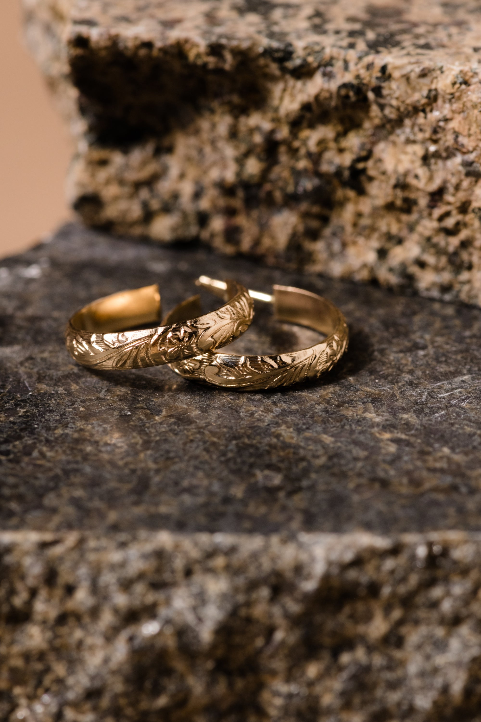 Close-up of two Filigree Hoops crafted in 14k gold-filled material, showcasing intricate leaf engravings, intertwined and resting on a rough, textured stone surface. The background features natural stone patterns that enhance the elegance of the hoops' floral design, beautifully contrasting with their shiny and detailed craftsmanship.