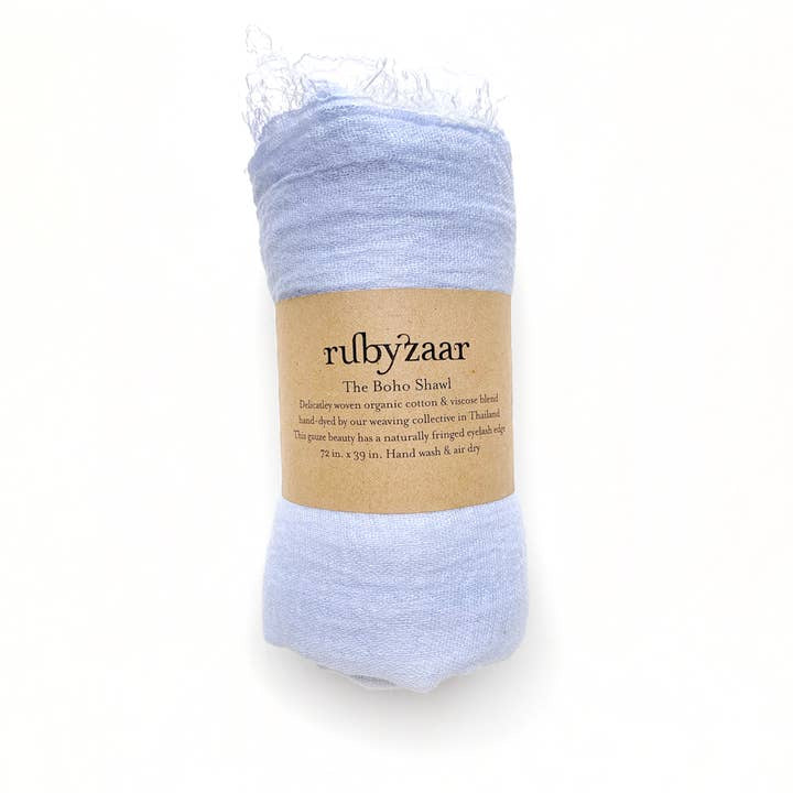 A light blue shawl, wrapped neatly with a kraft paper label, reads "rubyzaar Hima Large Scarf, Wrap, Shawl," describing the fair trade product as handwoven from organic cotton in Thailand. Measuring 72 x 39 inches, it's recommended for hand washing and air drying, offering an eco-friendly alternative to cashmere scarves.