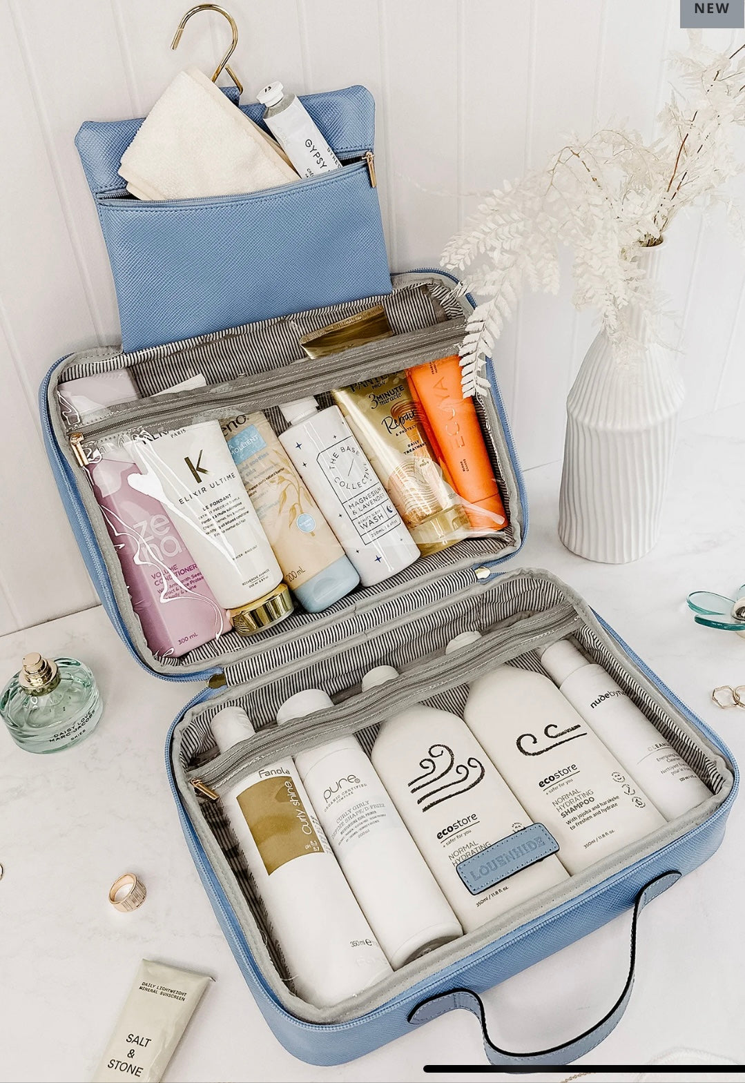 A blue Emma/Baby Audrey cosmetic bag from the set of 2 hangs open on a wall, showcasing its contents. The top compartment holds smaller items like tubes and a pouch, while the bottom compartment houses larger bottles. Beside it, a white vase with dried flowers is displayed, and assorted small beauty products are arranged on the marble surface.