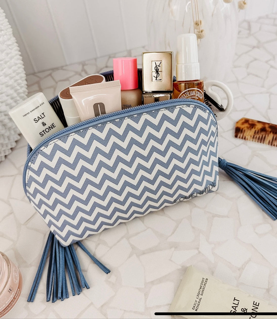 An Emma/Baby Audrey Cosmetic Bag from the set of two, featuring a blue and white chevron pattern with tassels, is open to display various beauty products. Nearby, you can see a brush, a small pink jar, and a white tube labeled "SALT & STONE," all arranged on a white mosaic-tiled surface.