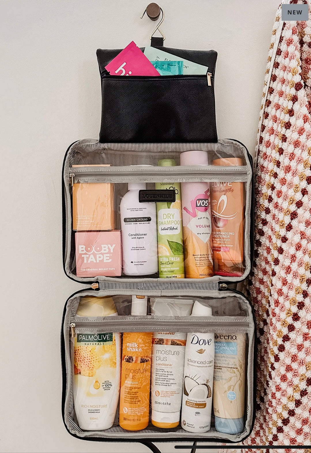The Emma/Baby Audrey Cosmetic Bags, featuring a sleek black exterior and a striped interior, make the ideal beauty case. They are perfect for organizing items such as conditioner, dry shampoo, VO5 volumizer, baby tape, and an assortment of lotions and face masks. Nearby hangs a colorful towel, while the top compartment unzips to store sanitary products conveniently.