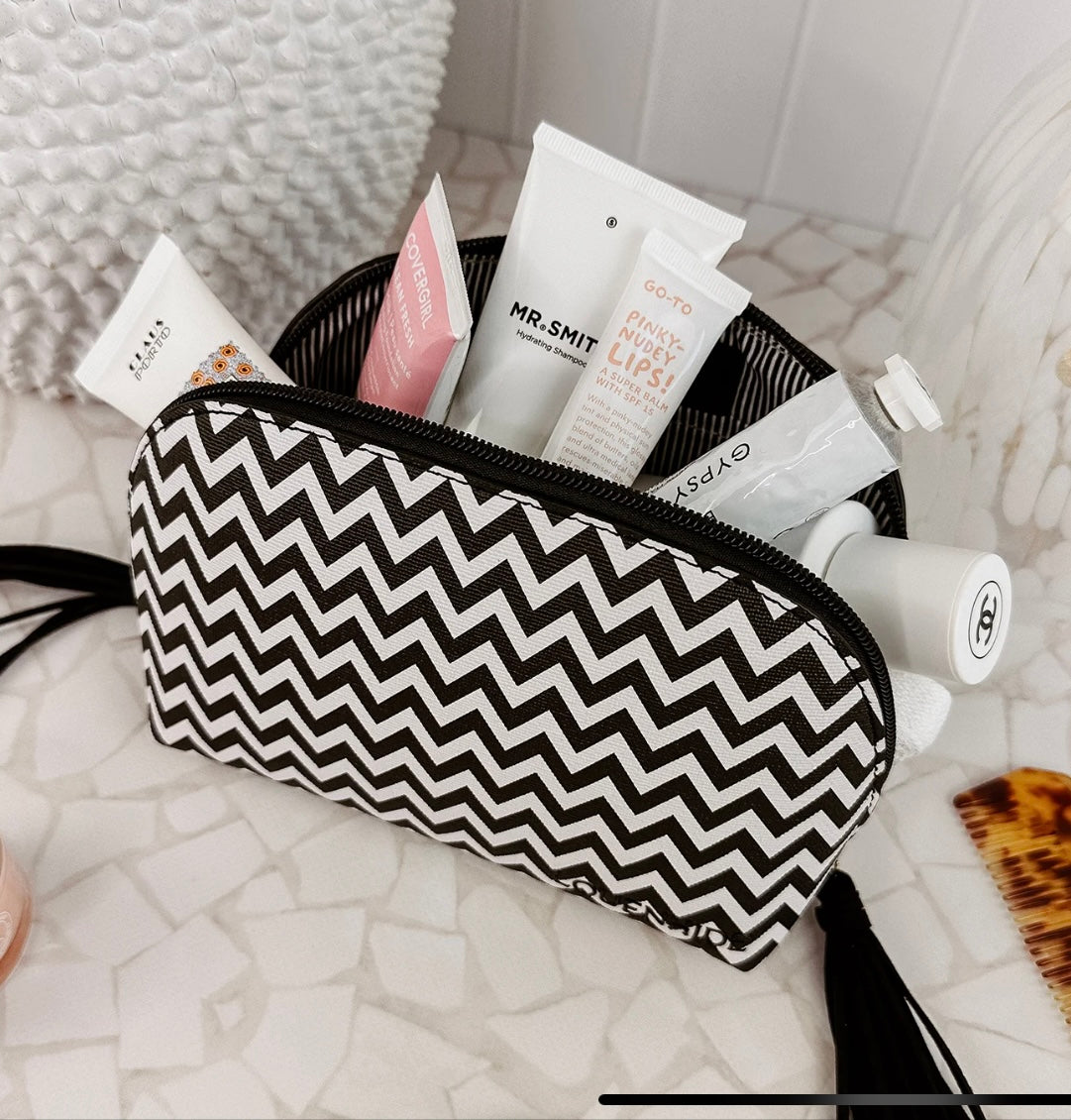 An Emma/Baby Audrey cosmetic bag from the set of two, featuring a black and white chevron pattern, is open on a tiled surface and filled with an array of skincare and beauty products. Visible items include tubes of cream, lip balm, and a bottle with a gold cap. A woven white basket is seen in the background.