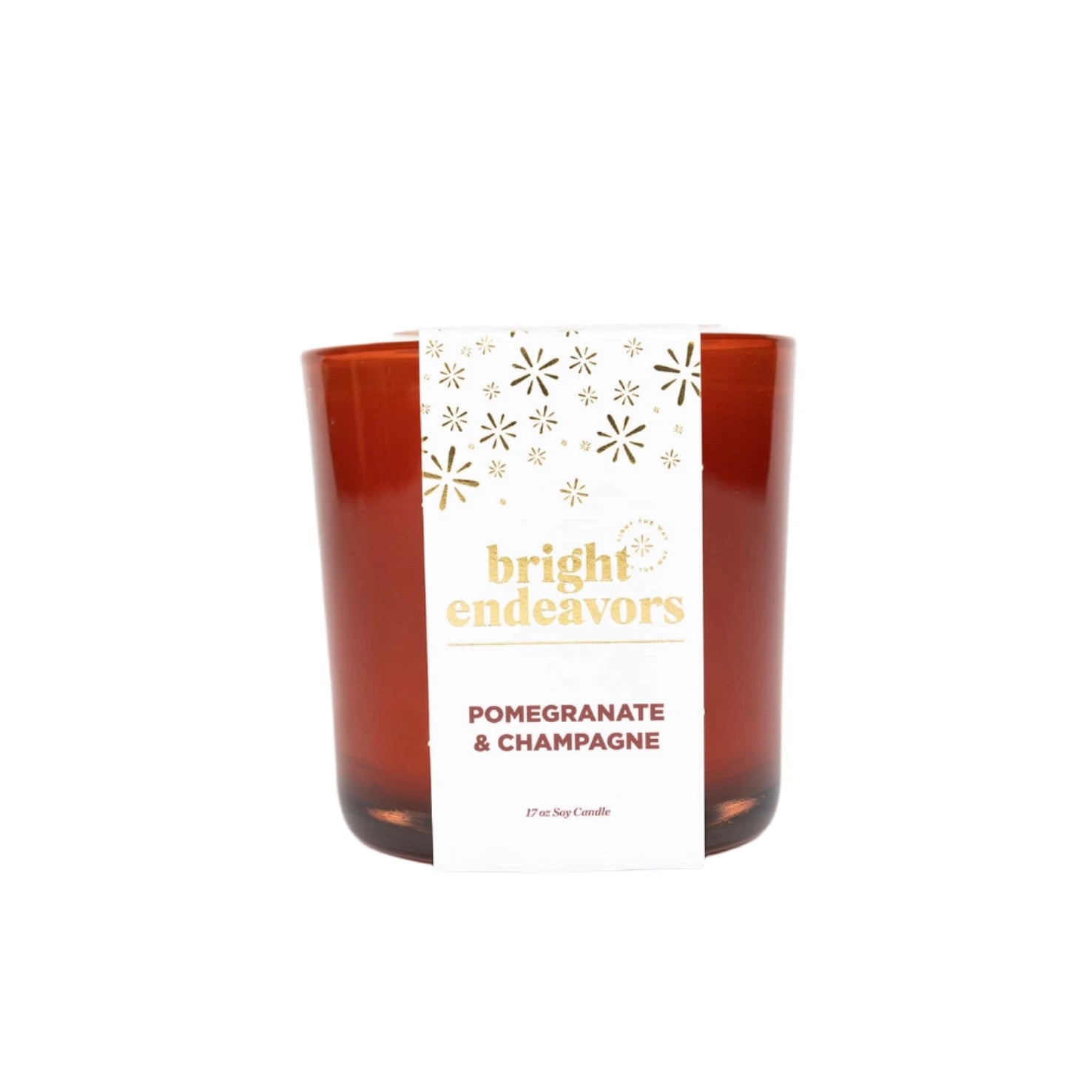 The Pomegranate & Champagne Soy Candle is a red glass candle with a white label adorned with gold text that reads "bright endeavors" and "POMEGRANATE & CHAMPAGNE." The label also includes elegant golden starburst designs. This 11 oz candle is made from soy wax and offers a phthalate-free fragrance, set against a plain white background.