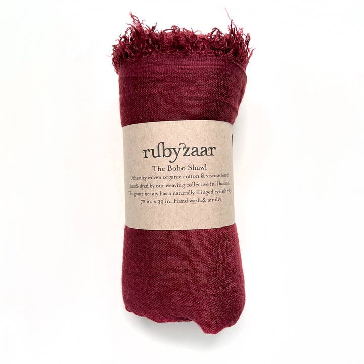 Rolled red scarf with fringe, wrapped in a brown label that reads "rubyzaar The Boho Scarf." This bohemian style accessory is hand-woven from a cotton viscose blend and hand-dyed by a weaving collective in Thailand. Dimensions are 72 inches by 29 inches. Care instructions include hand washing and air drying.