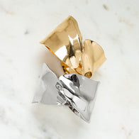 A sculptural statement ring showcasing two intertwined metallic bows, one gold and one silver, is elegantly displayed on a white marble surface. The crinkled, reflective texture mirrors the artistry of the Overlap Hammered Cuff, imparting a modern and artistic flair to the piece.