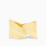 Introducing the Overlap Hammered Cuff: a stunning piece crafted from a golden, rippled sculptural form. This cuff showcases flowing curves and a central fold design, with its metallic texture shimmering beautifully against a plain white background, highlighting the artistry and brilliance of its contours.
