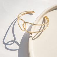 A Gold Twist Cuff with a 6" circumference rests on a white, round surface. The minimalistic, intertwined design of the bangle casts a distinct shadow. The light creates a soft, elegant atmosphere, highlighting its smooth texture and shiny finish.