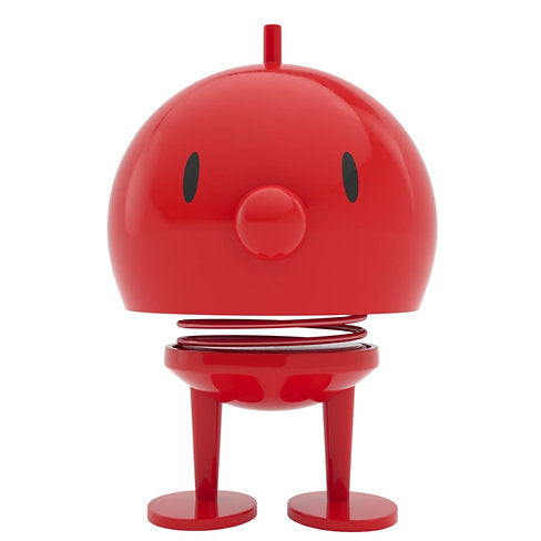 The Hoptimist Bumble is a red, round figure featuring a spring body, oval eyes, and a small nose. It stands on two flat feet and has an antenna-like top. Its simple yet playful design draws inspiration from Danish design history, offering both minimalist and cartoonish charm that makes it ideal for home decor.