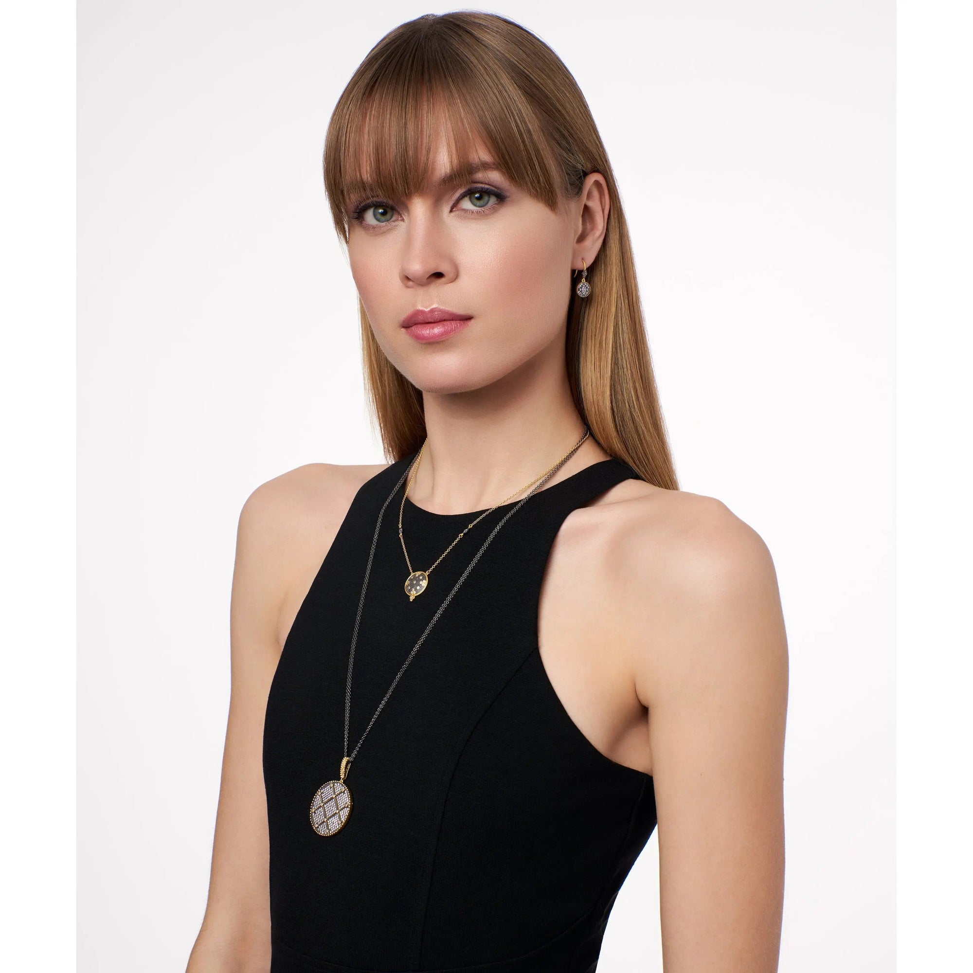 A woman with long, straight blonde hair and blunt bangs wears a sleeveless black dress. She accessorizes with two necklaces and small earrings. The longer necklace, the Signature Double Sided Pendant Necklace in sterling silver, features a large circular pendant adorned with a geometric pattern and cubic zirconia details. She stands against a plain white background.