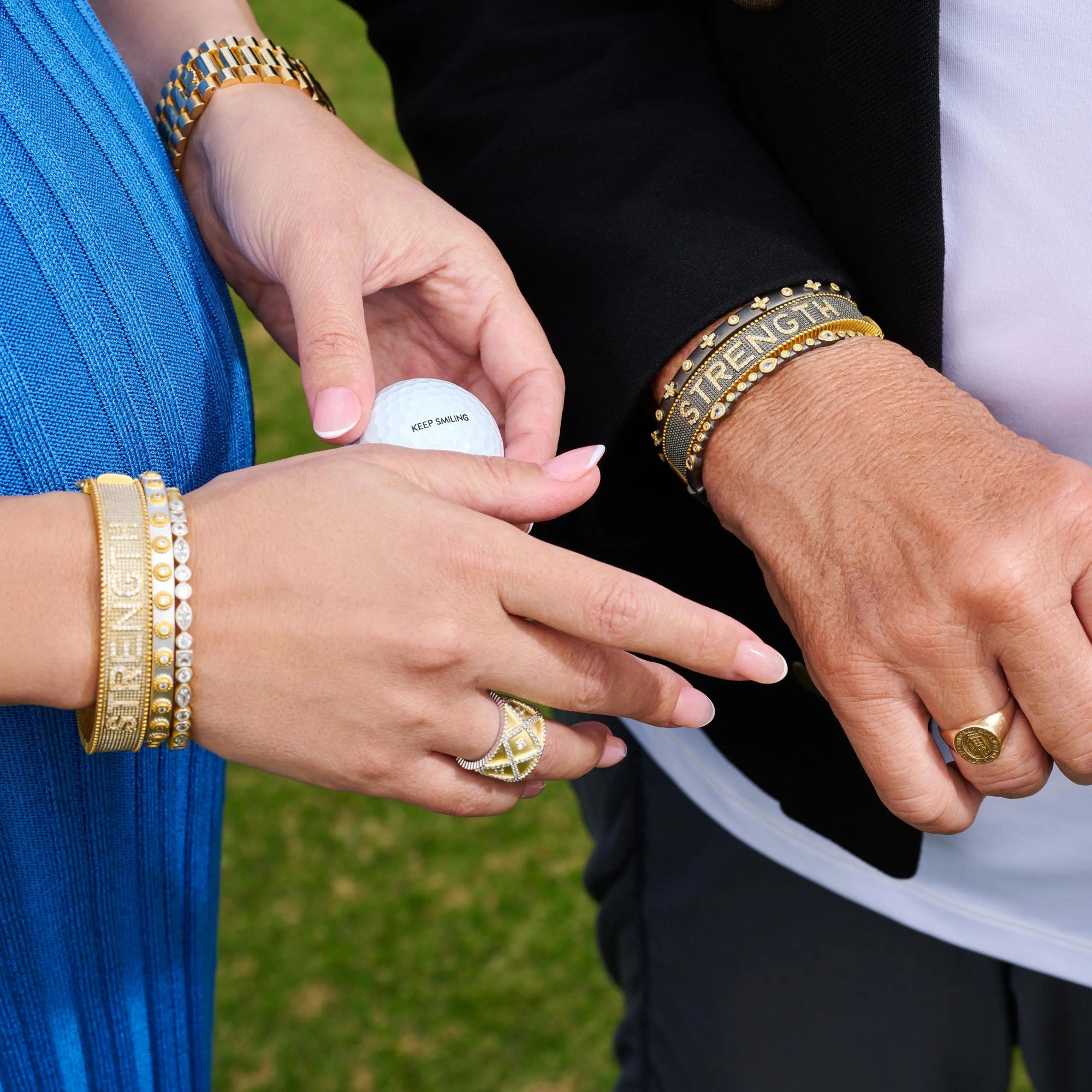 Two individuals with intertwined hands showcase The STRENGTH Bracelet, crafted in sterling silver and embellished with pavé stones. One person complements the bracelet with a gold watch, while the other wears a jeweled version of The STRENGTH Bracelet alongside a gold ring. Together, they hold a golf ball inscribed with "KEEP SMILING," all set against a lush grassy scene.