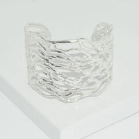 Displayed on a white surface is the Openwork Cuff, a wide, sculpted silver bracelet featuring an intricate wave-like texture. Its reflective and polished 24 kt gold plating enhances its elegant and modern design, while offering an adjustable fit for comfort.