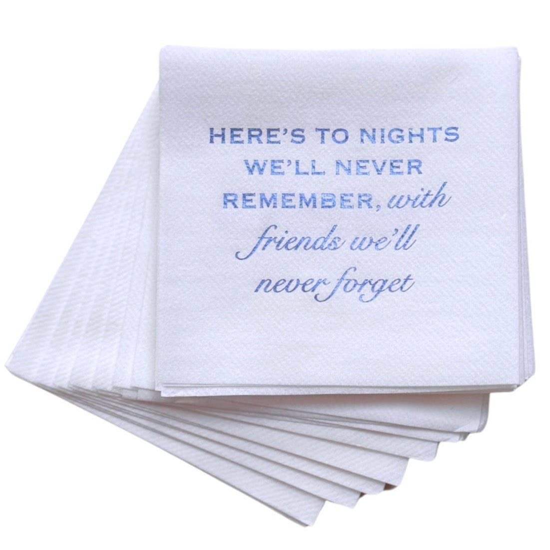 A stack of Year Round Entertaining Cloth-Like Cocktail Napkins in white features a foil embossed message: "Here's to nights we'll never remember, with friends we'll never forget." The napkins are neatly arranged, with one prominently showcasing the mix of uppercase and cursive fonts.