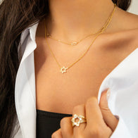 A close-up captures a person wearing a white shirt over a black top, showcased with gold-plated necklaces featuring the Star of David pendant and a small bar. A hand adorned with a star-shaped gold ring rests on the collar, partially open to reveal the layered jewelry.