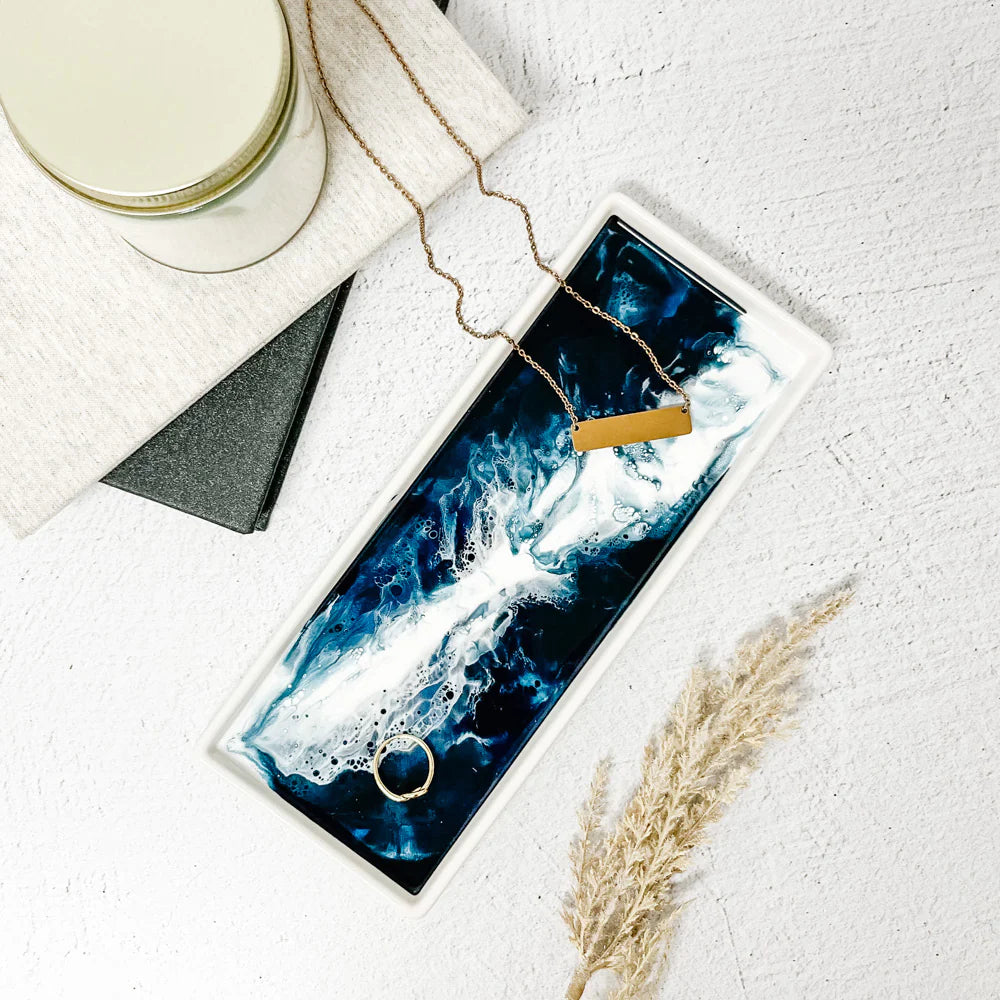 A Ceramic Resin Tray with a blue, white, and black marbled pattern is placed on a textured white surface. It holds a gold ring and necklace. Nearby, there is a closed candle, black notebook, white cloth, and dried wheat sprig, creating an elegant home decor arrangement.