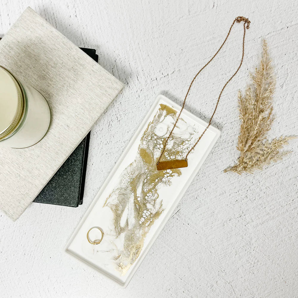 A *Ceramic Resin Tray* in white, with gold and white marbled patterns, elegantly holds a gold necklace and ring. It rests on a textured white surface. To the left, a candle sits atop a stack of beige-covered books, while dried pampas grass to the right adds to the home decor ambiance.