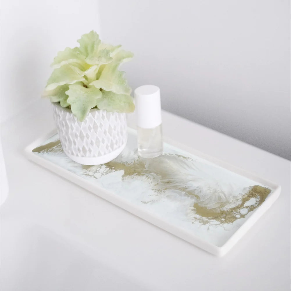 A Ceramic Resin Tray with a white and gold marbled design elegantly holds a small, textured white planter containing a leafy green plant, alongside a white nail polish bottle. The smooth, white background further complements the minimalist aesthetic of the home decor.