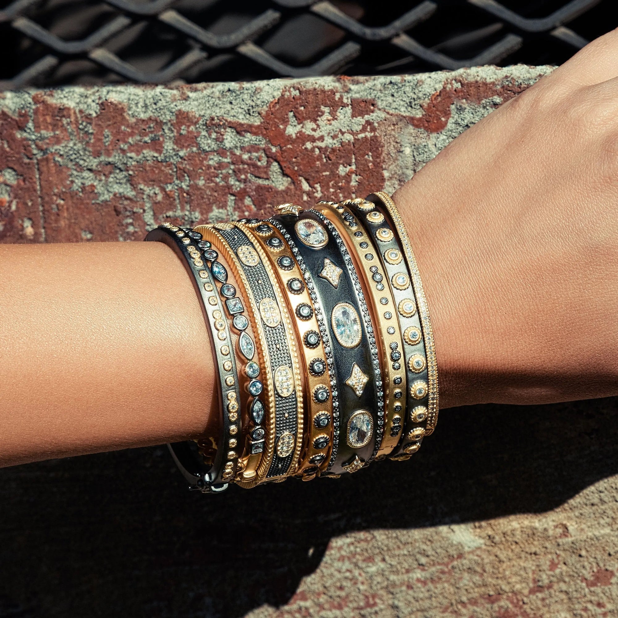 A close-up of a person's arm showcasing the Signature Mixed Shape Bangle, along with other bracelets in gold and black hues, including a 14K gold piece. The designs are detailed, featuring dots, diamonds, and cubic zirconia stones. The background is a textured concrete wall with an overlay of grid-like metal patterns.