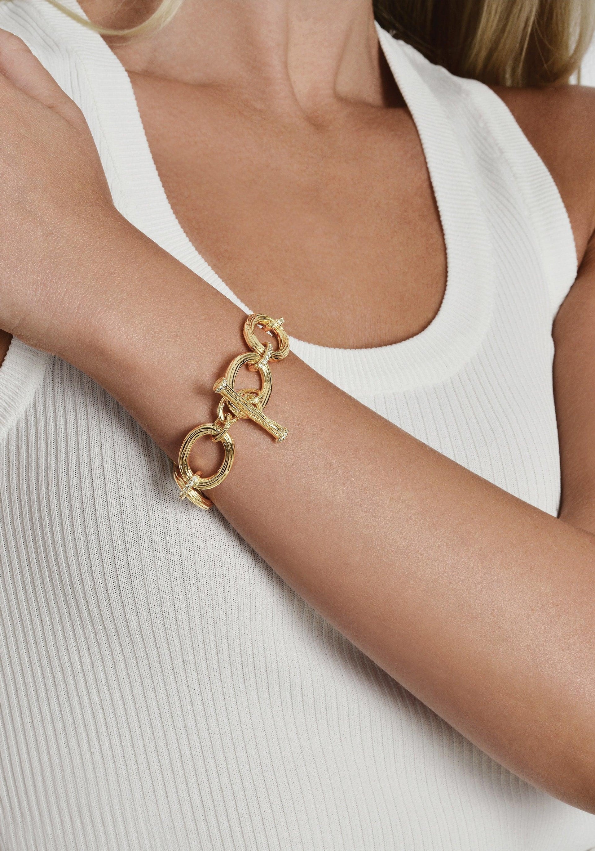 A person wearing a white ribbed tank top is spotlighting the stunning Enchanted Forest Chain Bracelet on their wrist, a chunky chain bracelet with large interlocking links crafted from gold-dipped brass. The individual's head and other hand are concealed, directing all attention to the captivating bracelet.