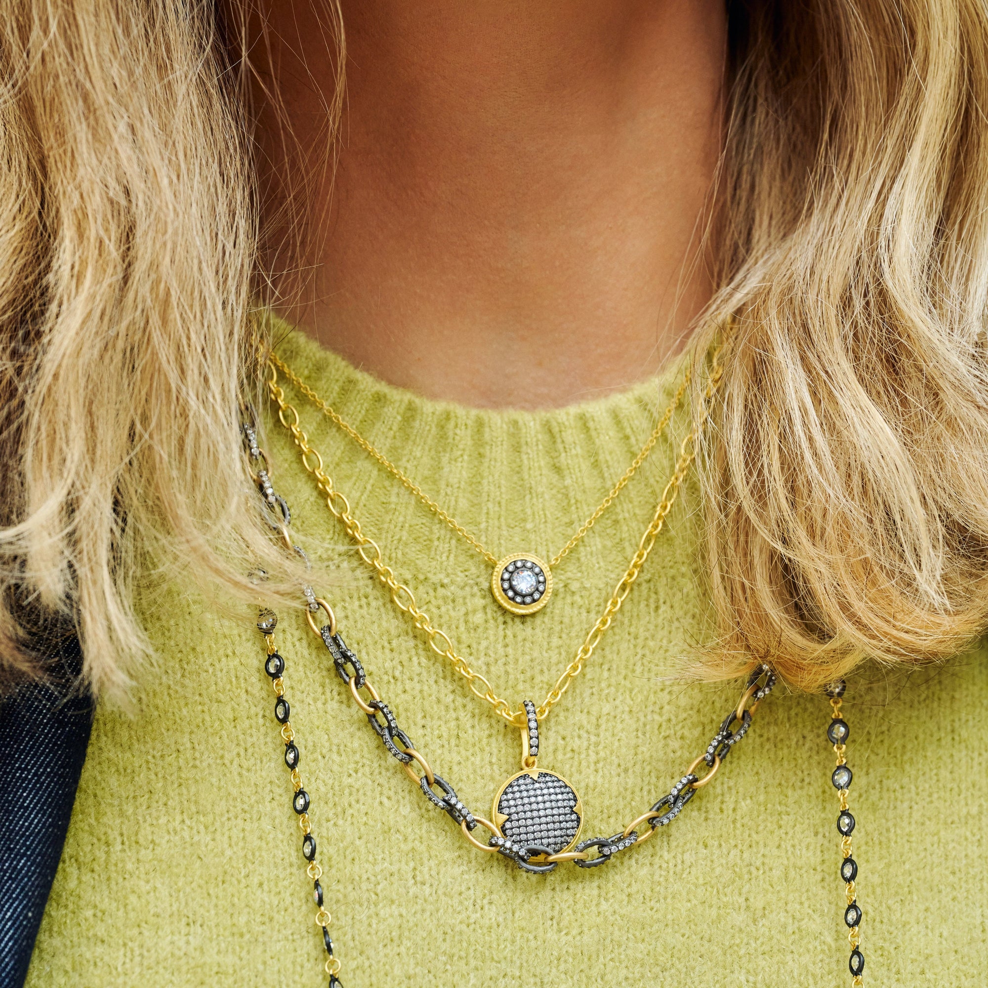 A woman is wearing multiple layered necklaces over a lime green sweater. Among them is the exquisite Signature Times Square Pave Short Pendant, crafted in 14K gold and adorned with intricate cubic zirconia designs. Her blond hair partially drapes over the shimmering gold and silver chains.