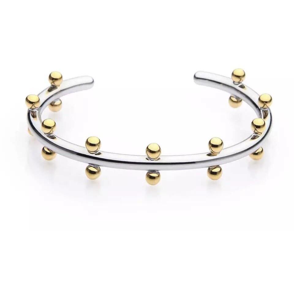 The Cindy Studded Cuff is an open silver bracelet featuring 18k gold-plated beads that are evenly spaced along its outer edge, creating a modern and elegant design. This hypoallergenic piece offers sophistication and is elegantly displayed against a plain white background.